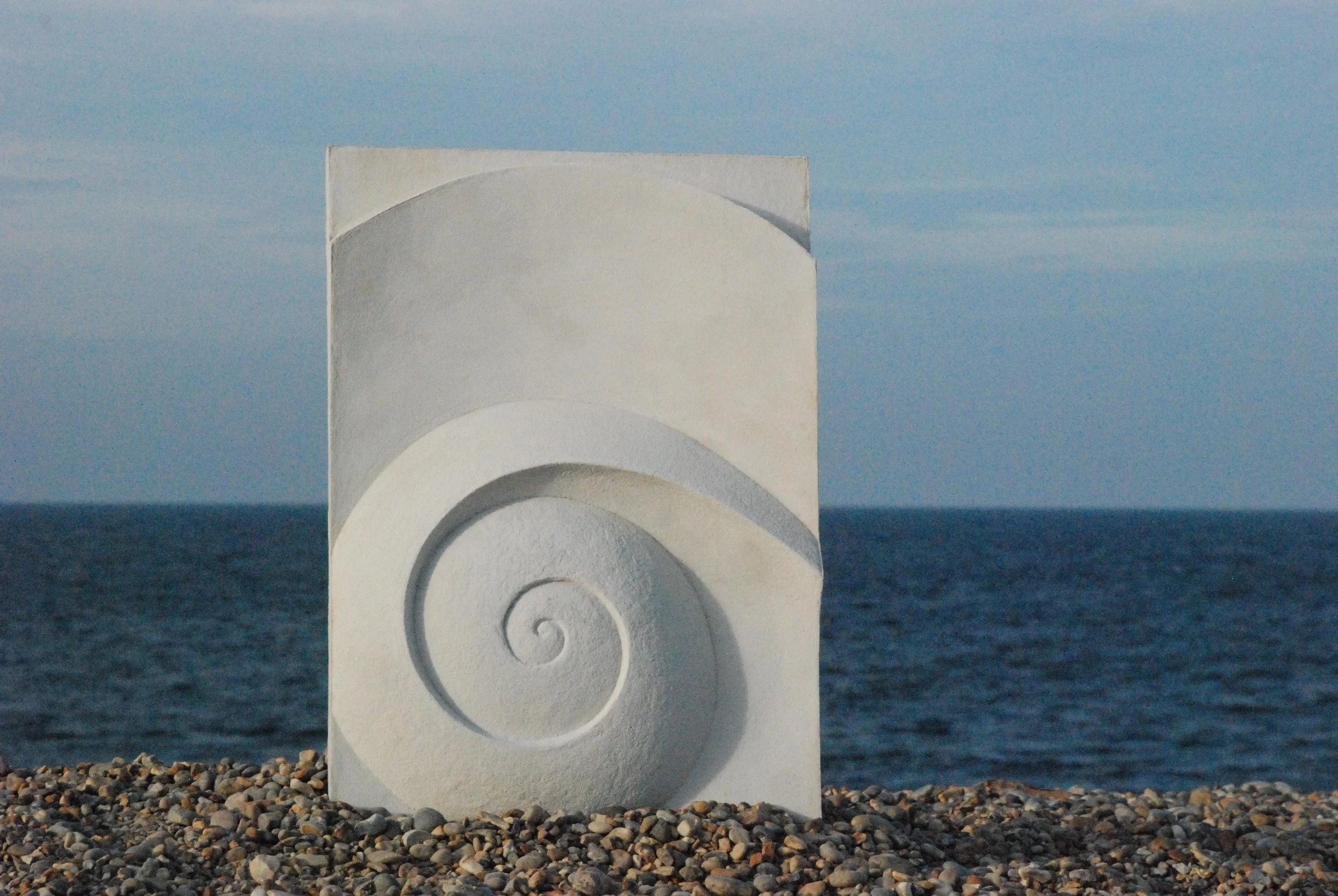 Moonspiral wall sculpture standing on a shingle beach with the sea and evening sky behind