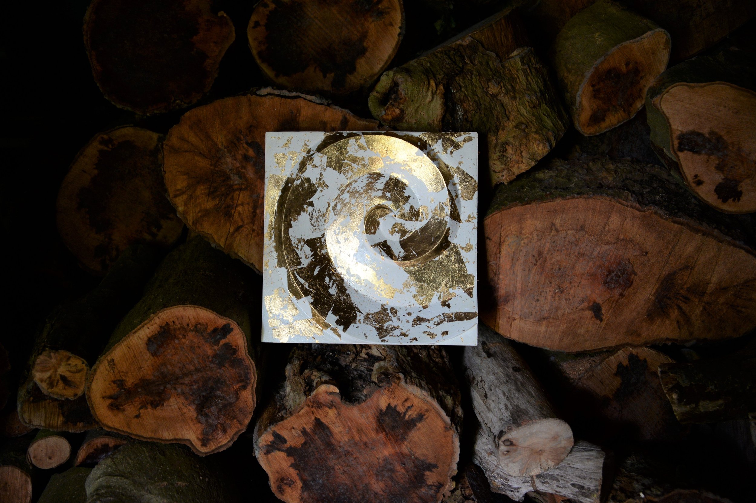 Golden Spiral wall sculpture, a small gold-leaf spiral panel displayed against a stack of cut logs