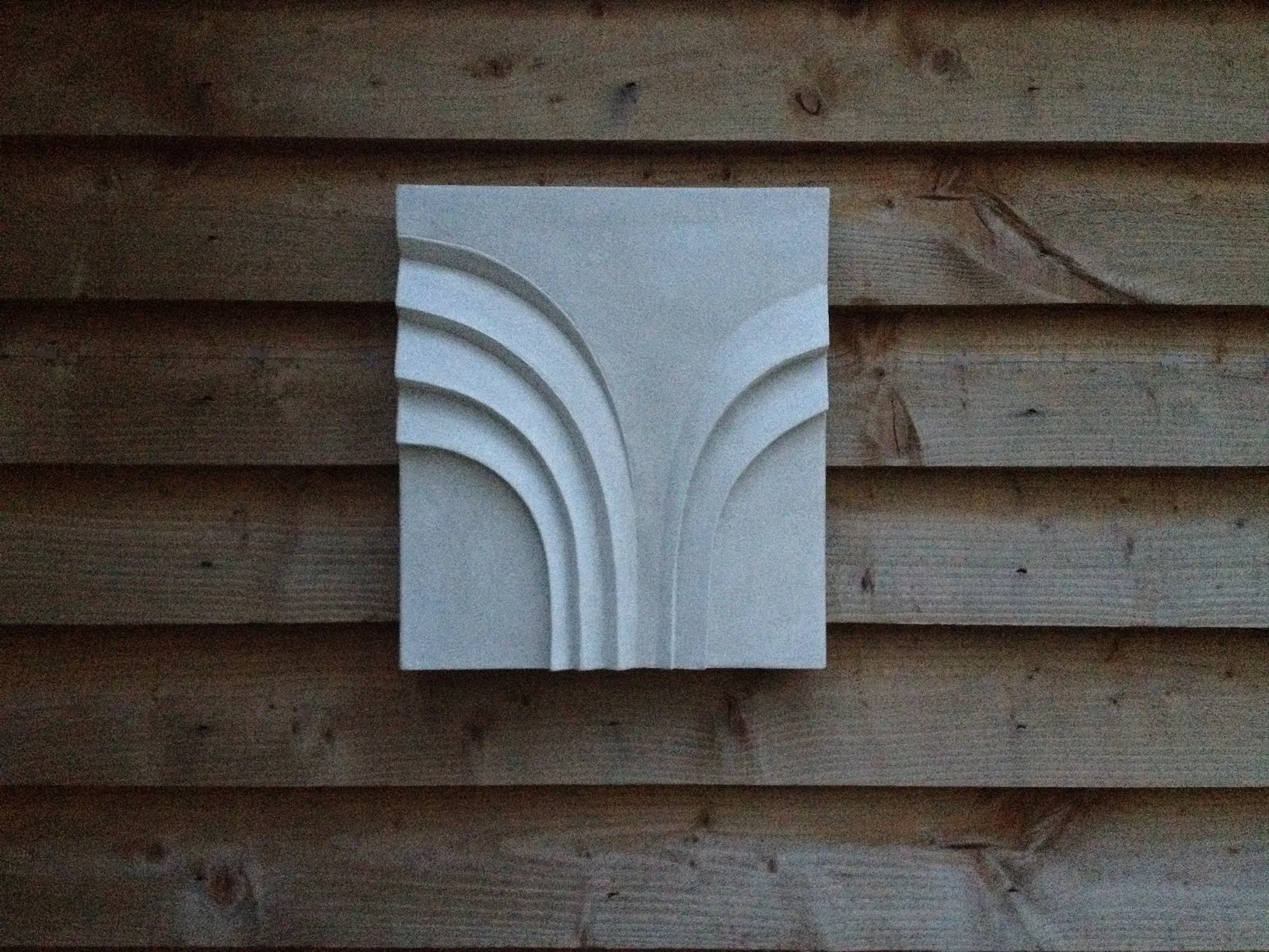 Palm Form Two wall sculpture, a pale stone-finish panel carved with a fanned palm frond relief, on timber cladding