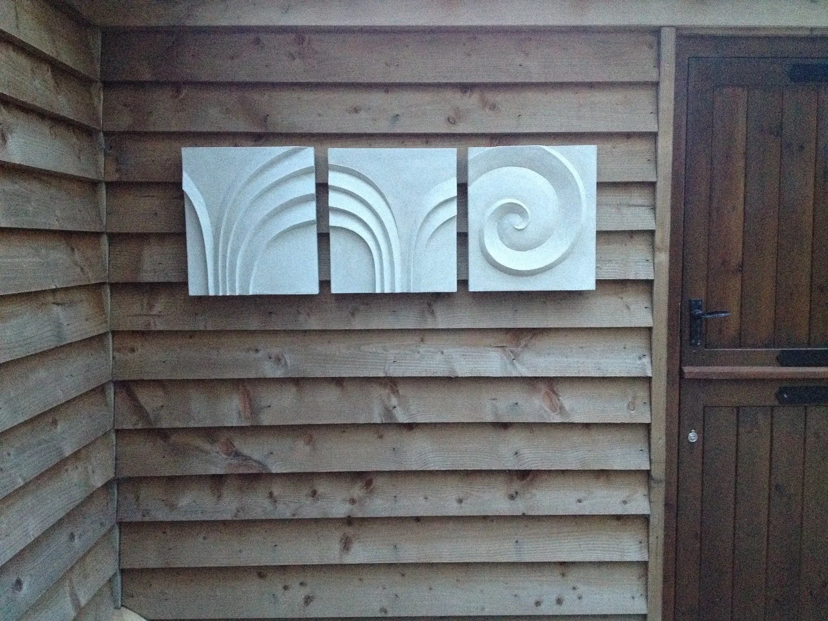 Two Palm Forms with Spiral wall sculpture, a three-part carved relief in pale stone finish on a timber clad wall