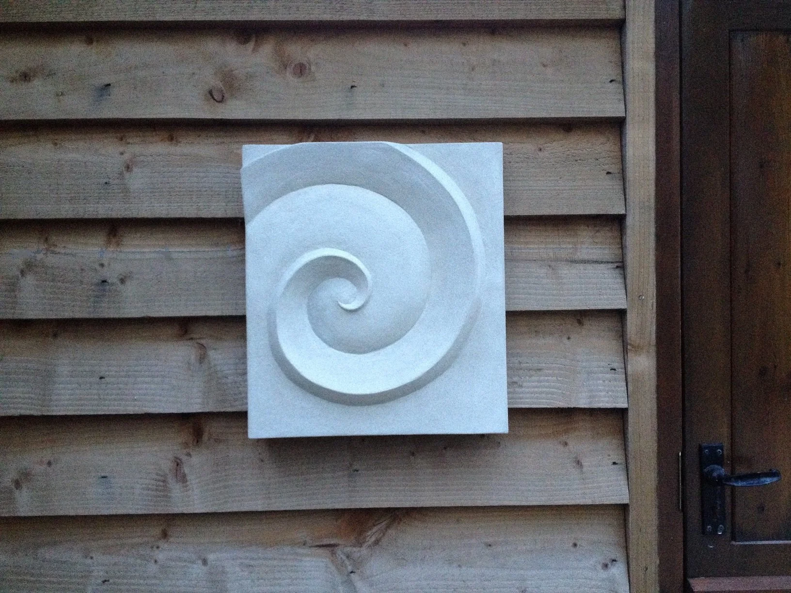 Spiral of Palm Form wall sculpture, a pale stone-finish panel carved with a single deep spiral, on timber cladding