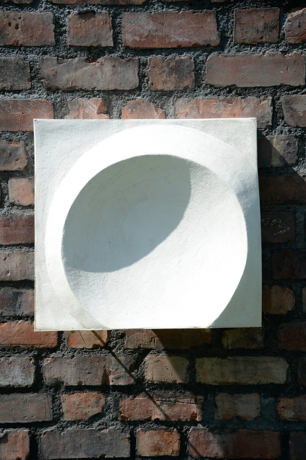 Moonwater wall sculpture, a 50cm concave lunar disc in white stone finish set in a square panel on a brick wall