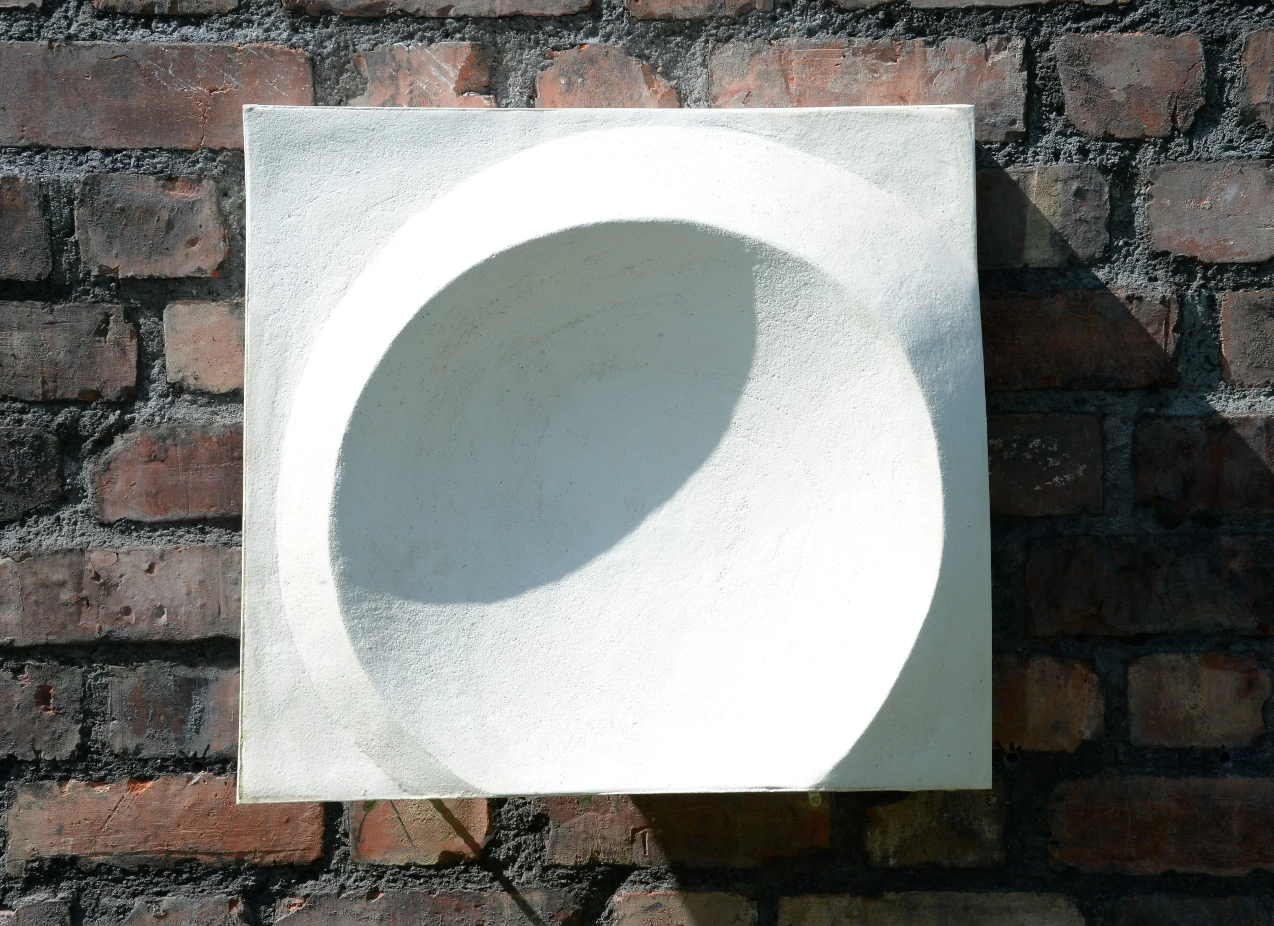 Moonwater Catcher wall sculpture, a white stone-finish square panel with a deep concave circular hollow, on a red brick wall