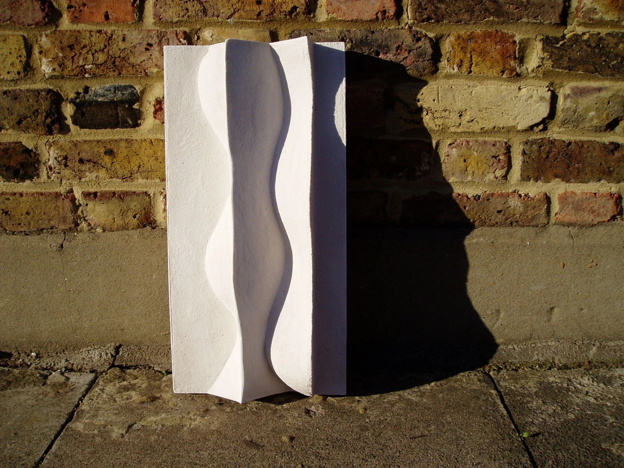 Tall white Brancusi 2 relief beside brickwork, with a strong shadow along its curved ridge.