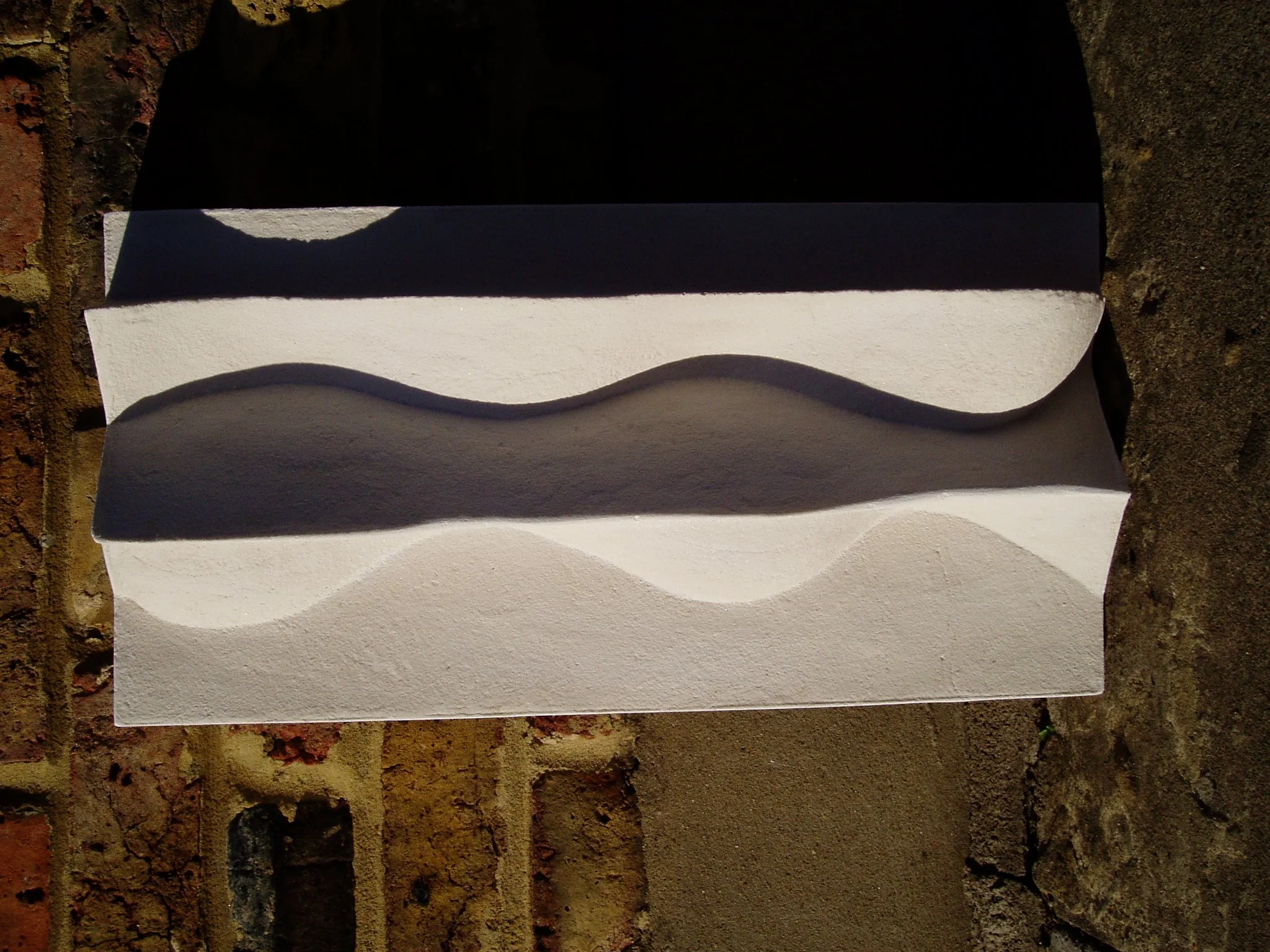 White Brancusi 2 relief displayed horizontally in strong light against brick.