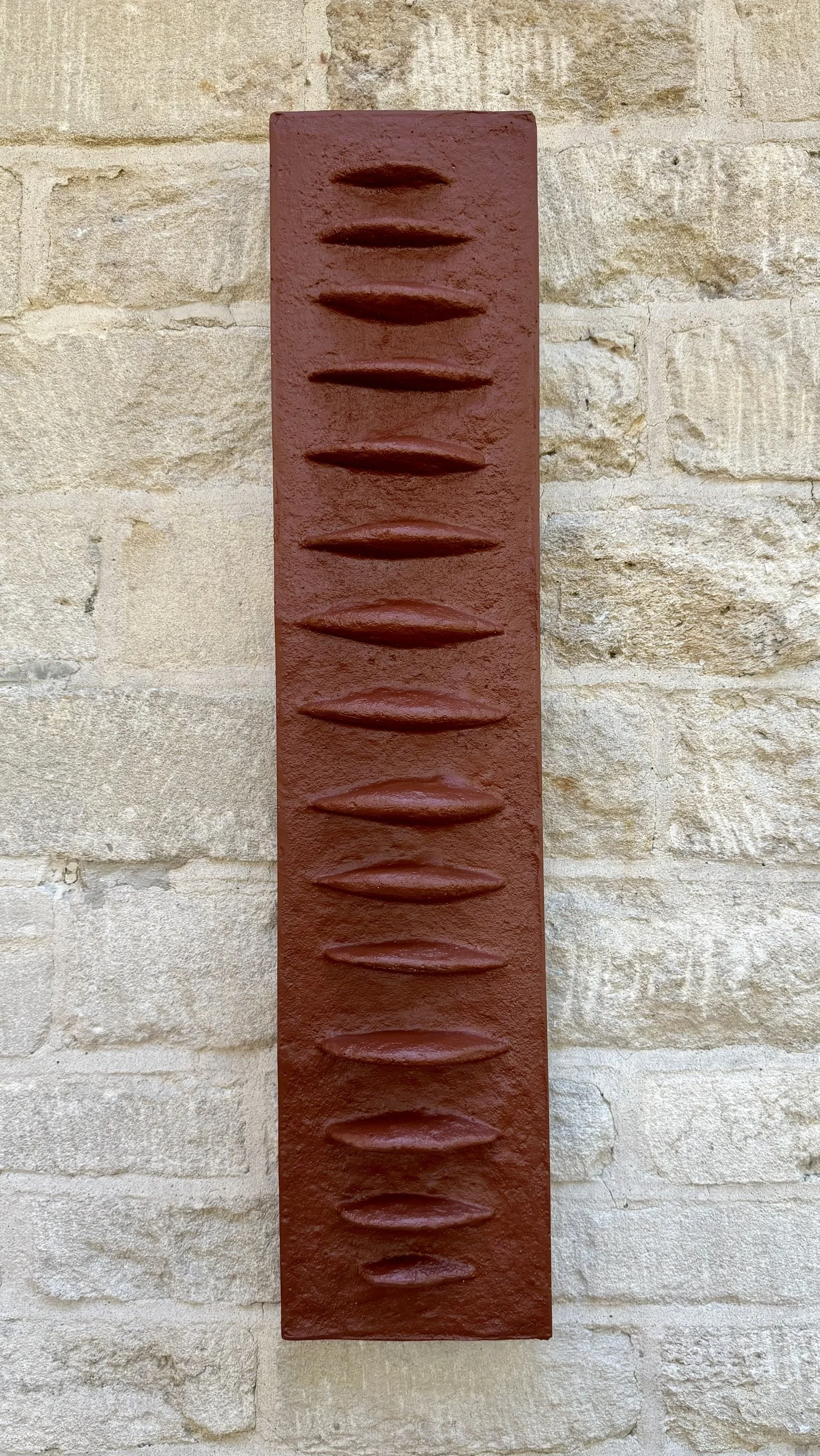 Terracotta Frozen Fish with a vertical row of raised fin-like forms on a stone wall.