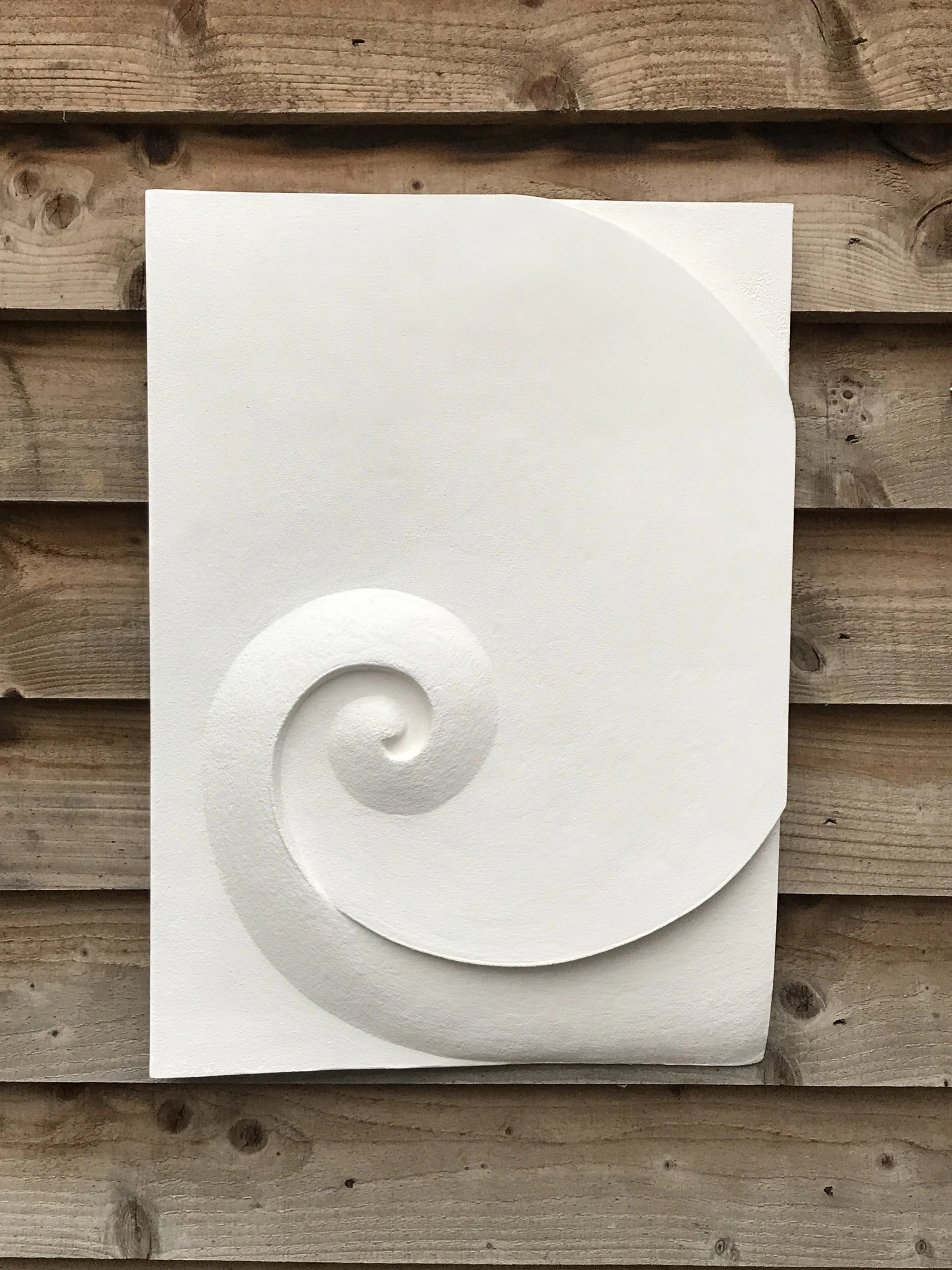 White Moonspiral 1 rectangular relief with a broad spiral, photographed against a wooden fence.