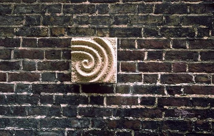 Wider view of the beige Raked Spiral 3 spiral relief against brickwork.