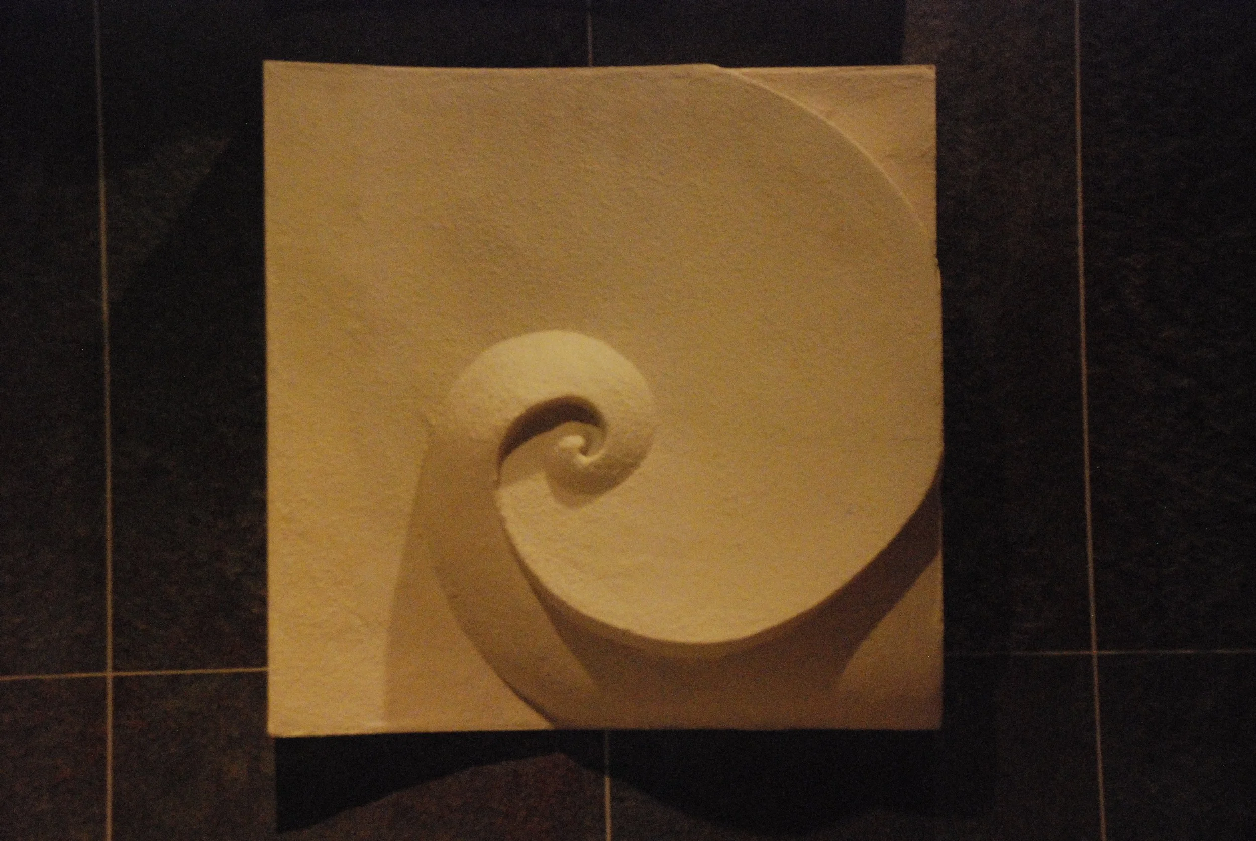 Wave and Moonshadow 1 spiral relief photographed in warm light against a dark tiled background.