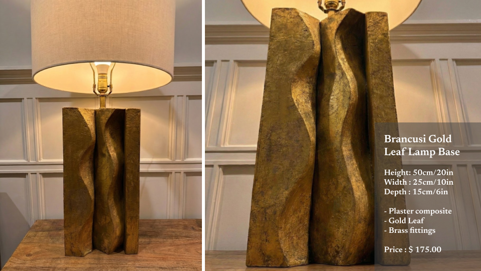 Brancusi Gold Leaf lamp base with a wavy, abstract design in gold leaf, placed on a wooden table with a beige lampshade on top, against a white paneled wall.
