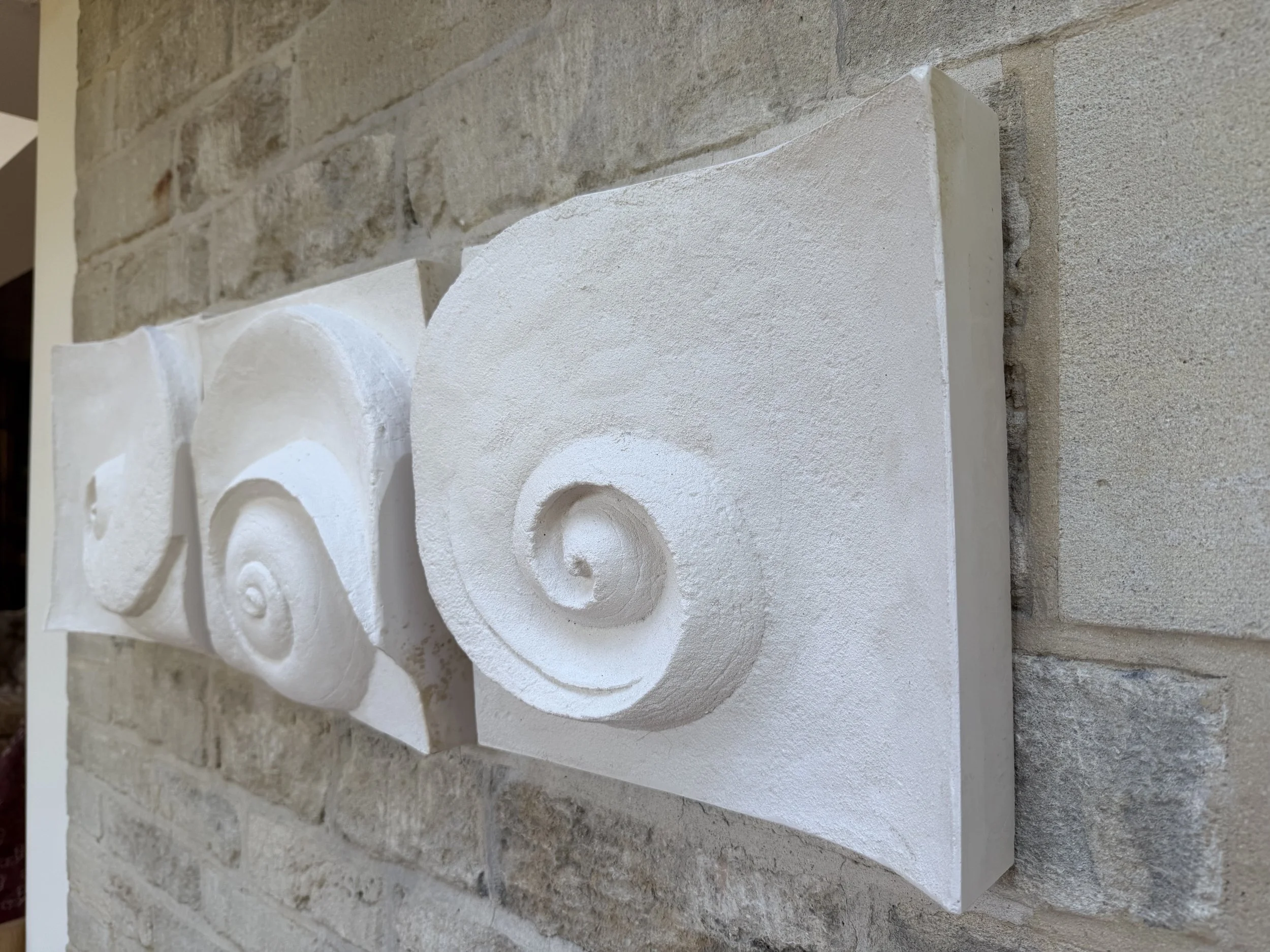 Three Wave Phase White panels seen in perspective along a stone wall, their spirals repeating