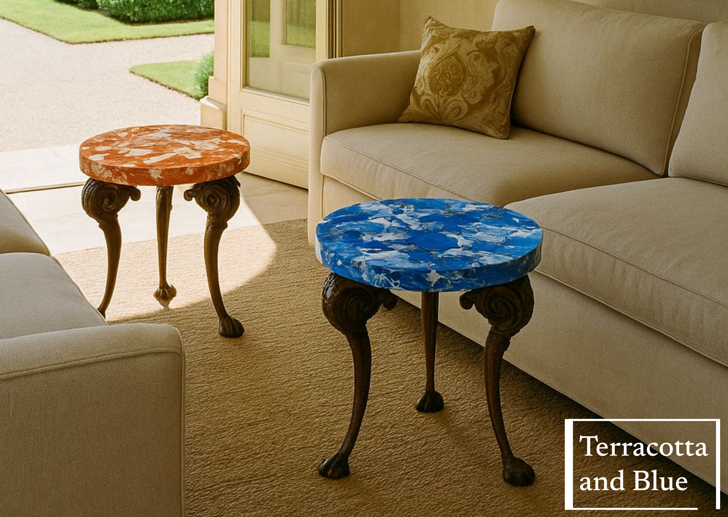 Two side tables with marble tops, one terracotta and one blue, with ornate wooden legs, placed between beige sofas in a sunlit room.