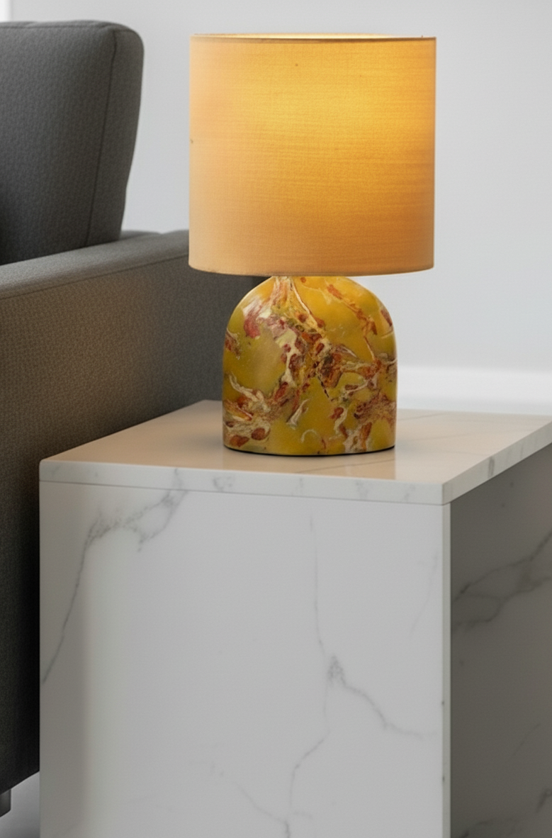 A table lamp with a yellowish-orange shade and a marble-like base on a white marble side table next to a gray upholstered sofa.
