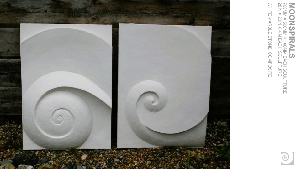 Two white stone sculptures with spiral and curved designs, placed outdoors against a wooden background.