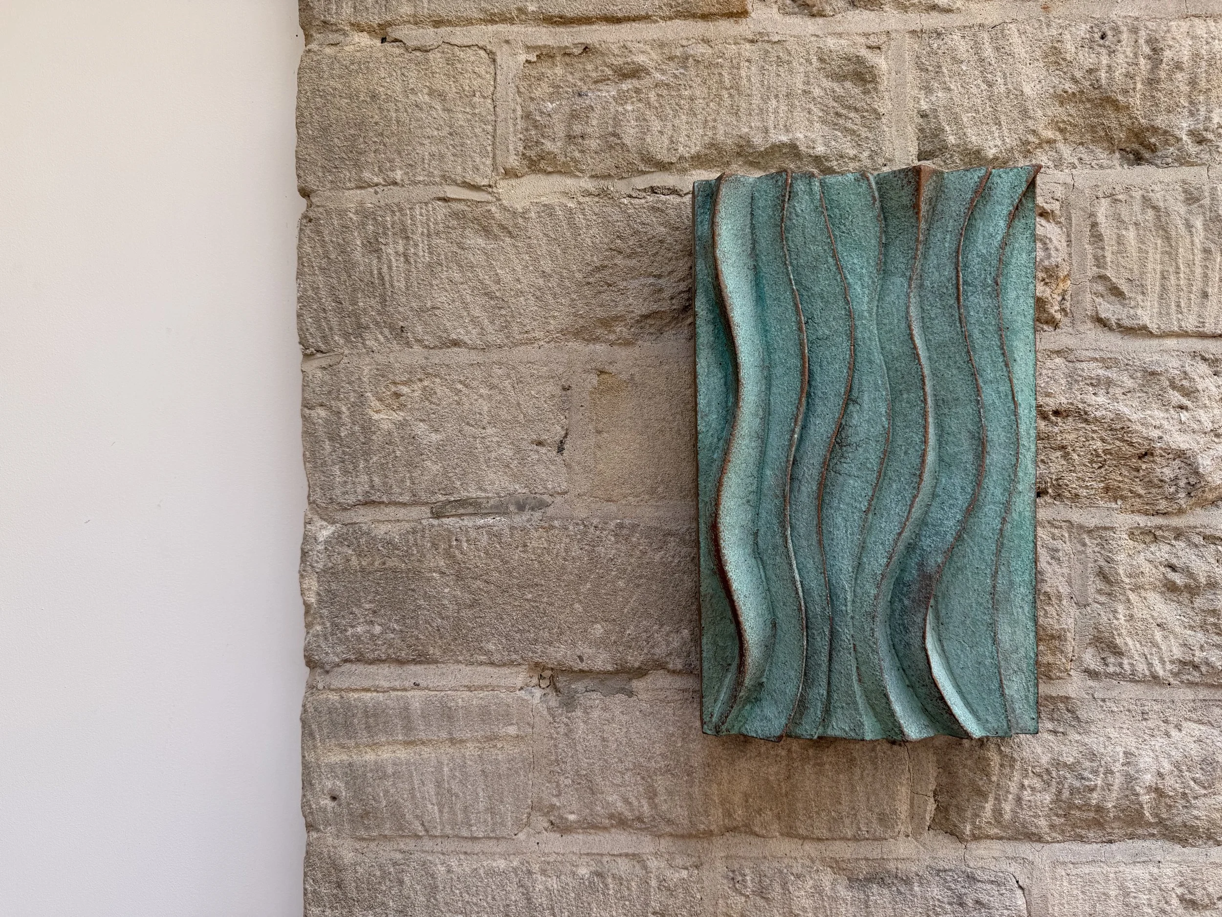 Verdigris Ripple Wave Wall Sculpture on a pale stone wall, the green rippled panel catching side light