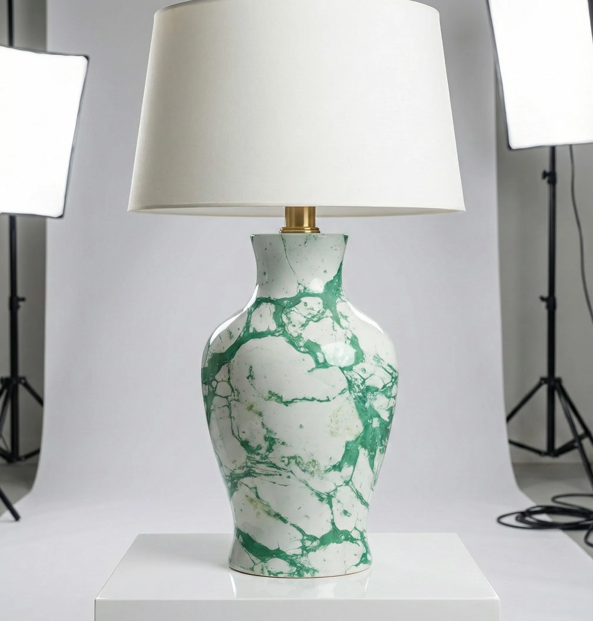 Ceramic table lamp with a white and green marbled pattern, topped with a white lampshade, on a white display stand, with photo studio lights in the background.