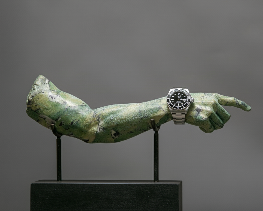 A sculpture of an outstretched arm with a wristwatch, mounted on a black stand against a gray background.