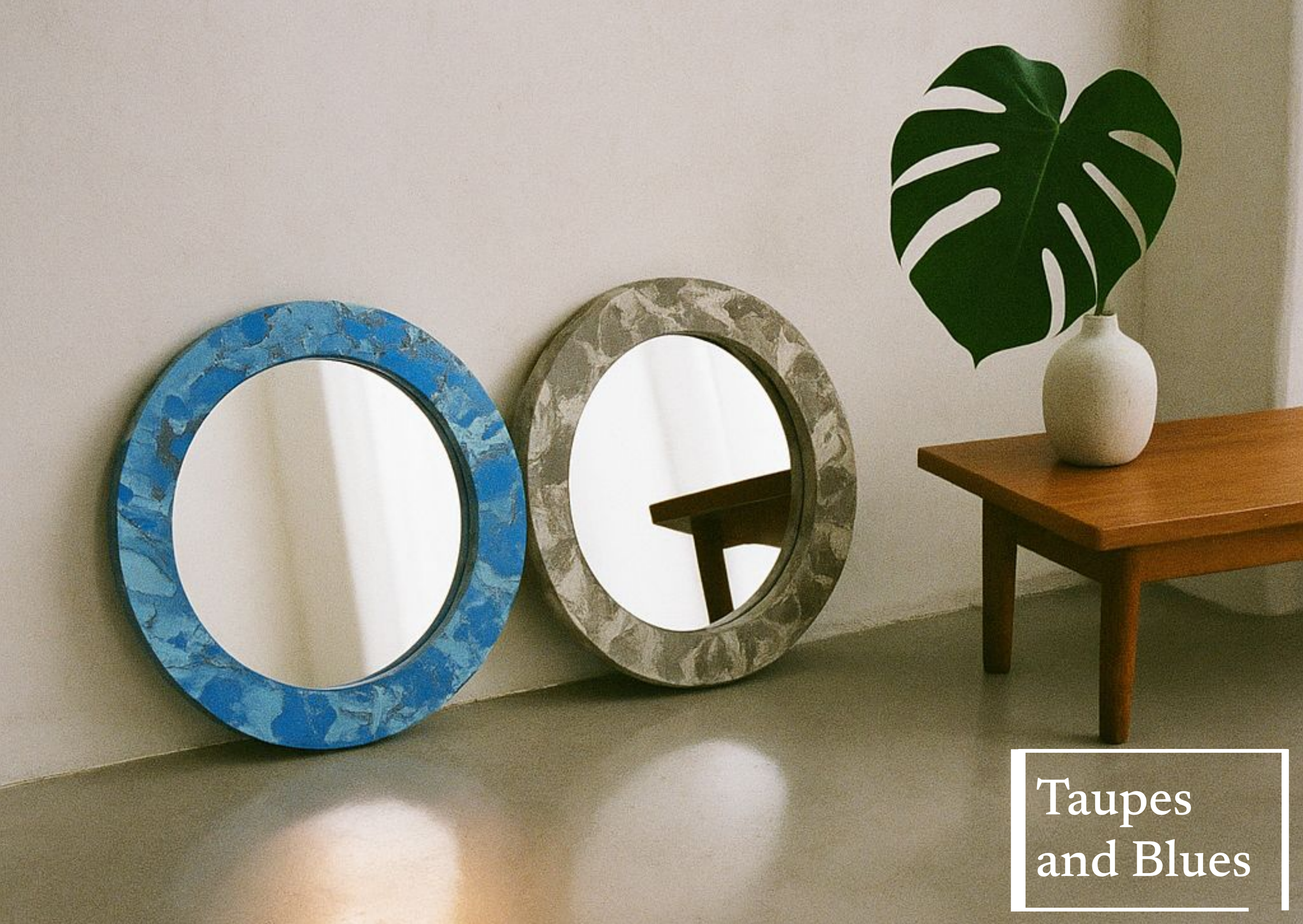 Two decorative round mirrors with blue and grey marble-like frames leaning against a white wall, on a light-colored floor. To the right, a wooden table with a white vase holding a large green Monstera leaf. Text overlay reading 'Taupes and Blues' in