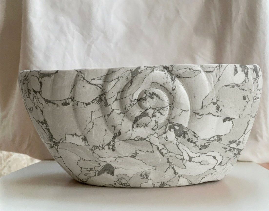 White marble sculpture with dark gray veins, featuring a shell-like spiral design in the center, placed on a white surface with a crumpled light-colored background.