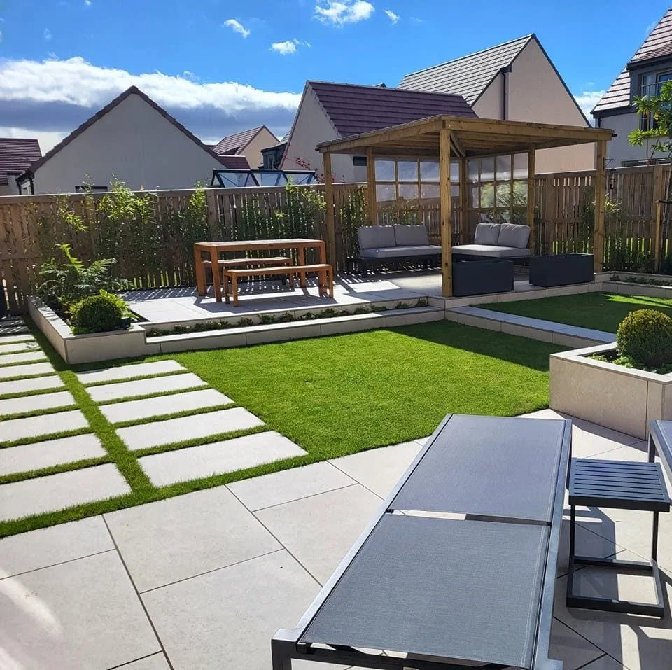 Backyard with a paved patio, lounge chair, small side table, grassy area, raised garden beds, a wooden gazebo with outdoor seating, and neighboring houses in the background under a blue sky with clouds.