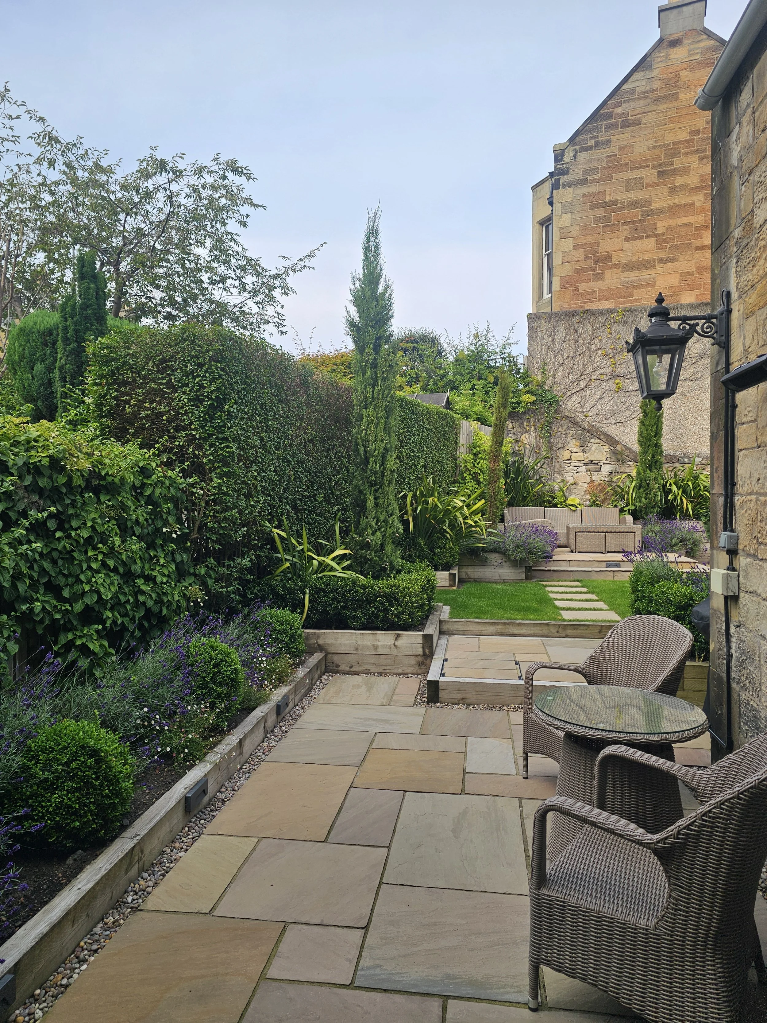 A backyard patio with stone flooring, surrounded by green shrubs and trees. There is a wicker chair and a small round glass table, with a larger stone building and a decorative lamp on the wall in the background.
