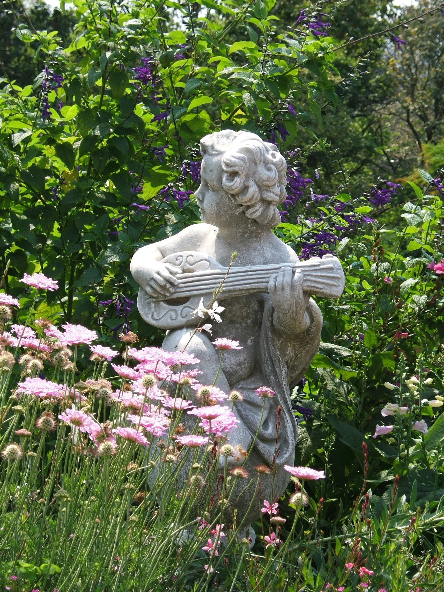 A stone sculpture of a woman holding a guitar, surrounded by pink and purple flowers and lush green foliage in a garden.
