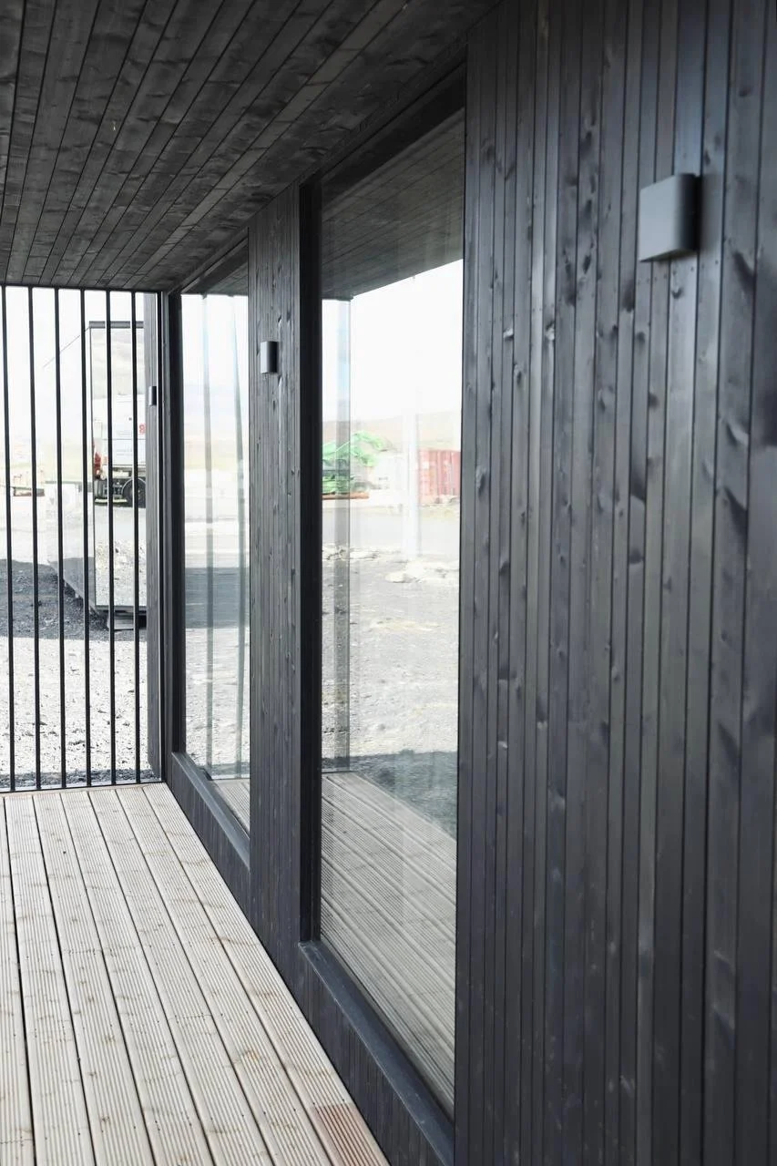 Interior view of a modern building with black wooden paneled walls and ceiling, large glass windows, and a wooden deck floor.
