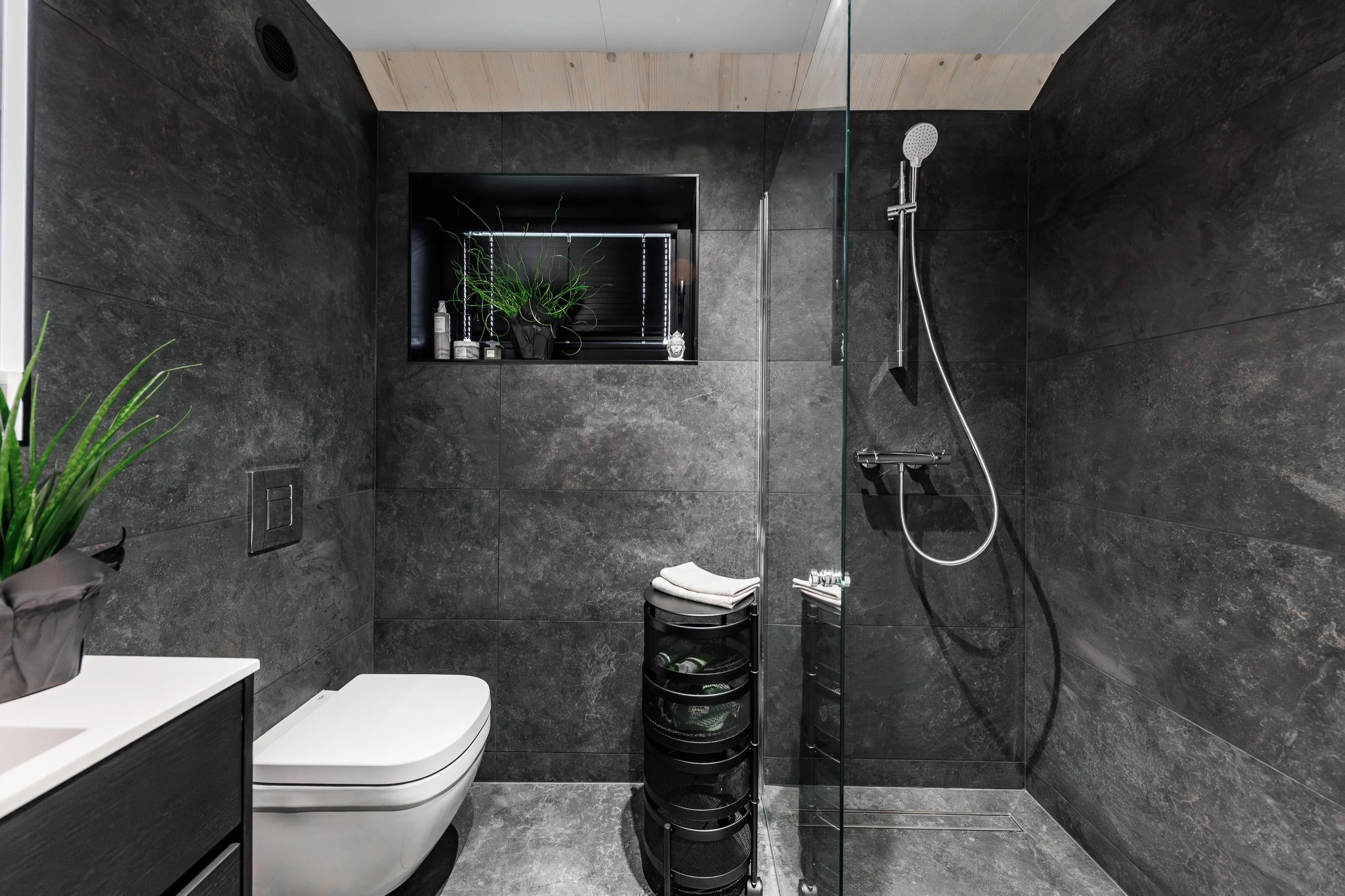 Modern bathroom with dark gray tiled walls and floor, a white toilet, a small black storage unit with towels, a small black window ledge with a plant and toiletries, and a walk-in shower with a glass panel, a silver showerhead, and a handheld shower