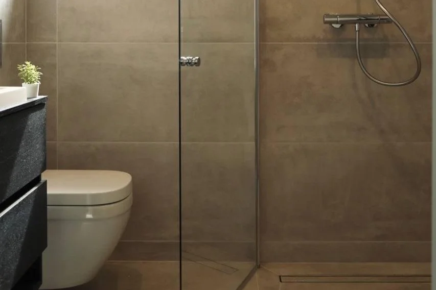 Modern bathroom with beige tiled walls, a white toilet, a black vanity with a small plant, and a glass-enclosed shower with a handheld showerhead.