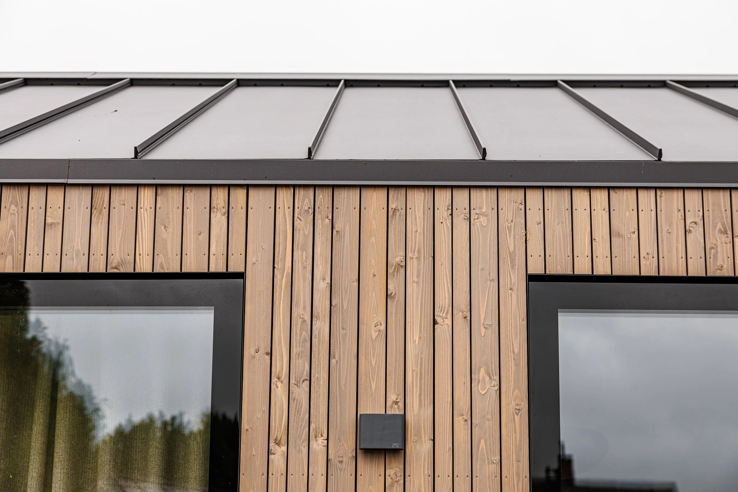Close-up of a modern building with vertical wooden siding, two large windows with black frames, and a section of a metal Ruukki roof with ridges.