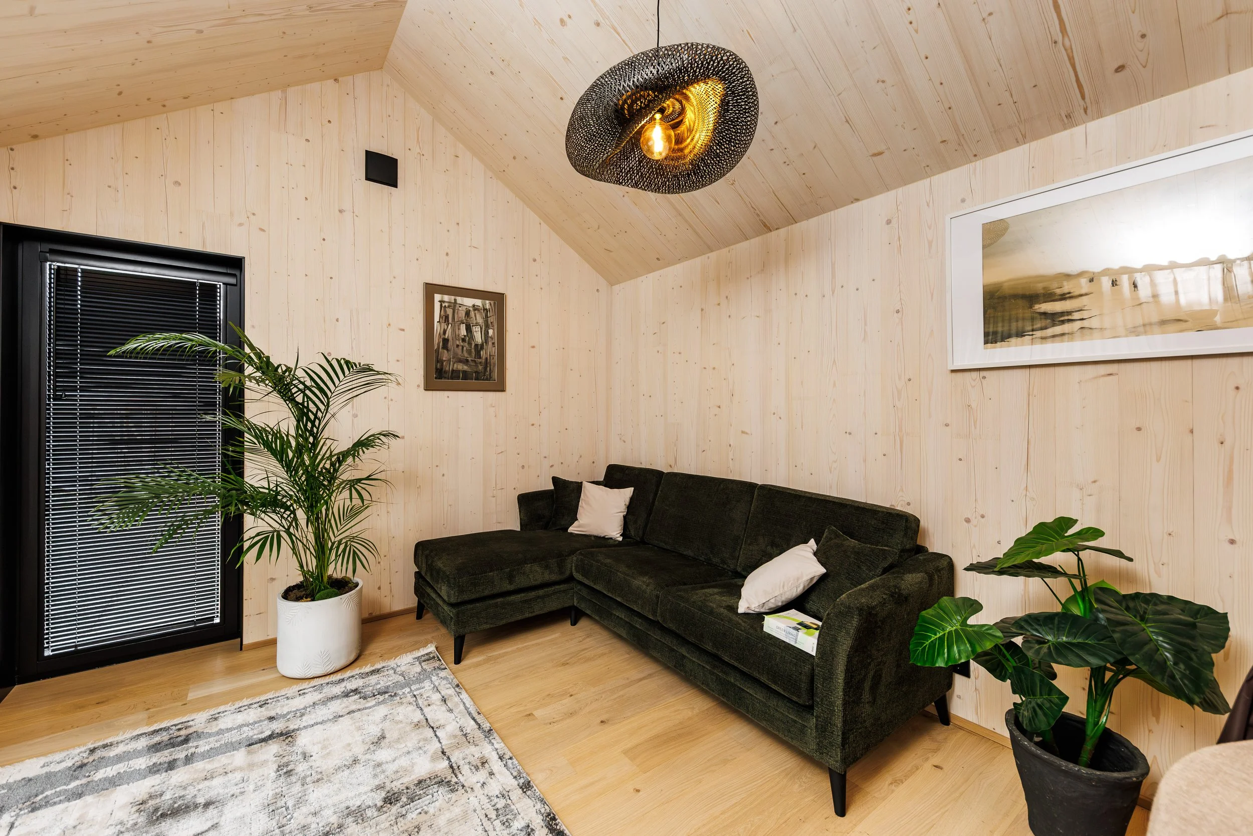 Living room with wood-paneled walls, a black sectional sofa with pillows, two potted plants, framed wall art, a window with blinds, and a modern light fixture on the ceiling.