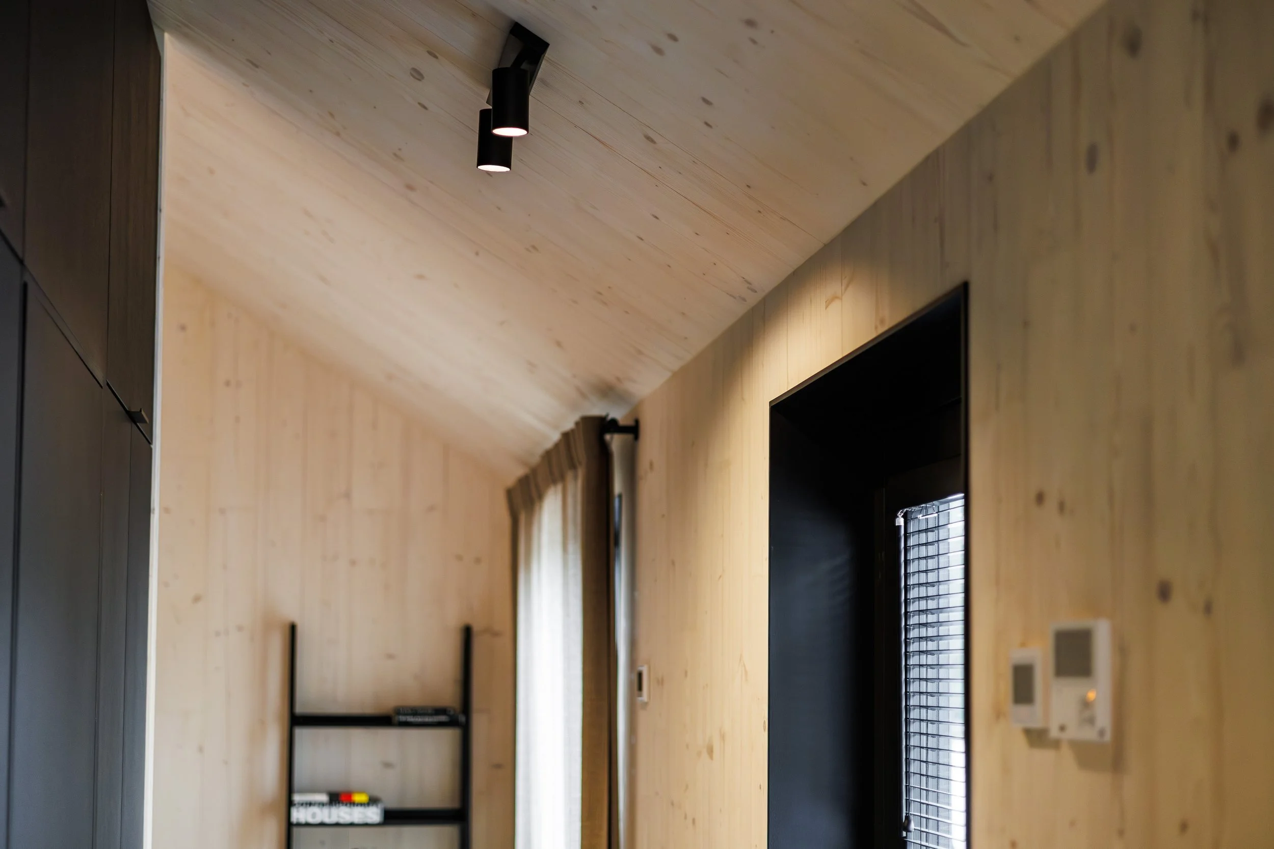 Interior view of a room with wood-paneled walls and ceiling, black ceiling lights, a window with curtains, a black door, and a black shelf with books and decor.