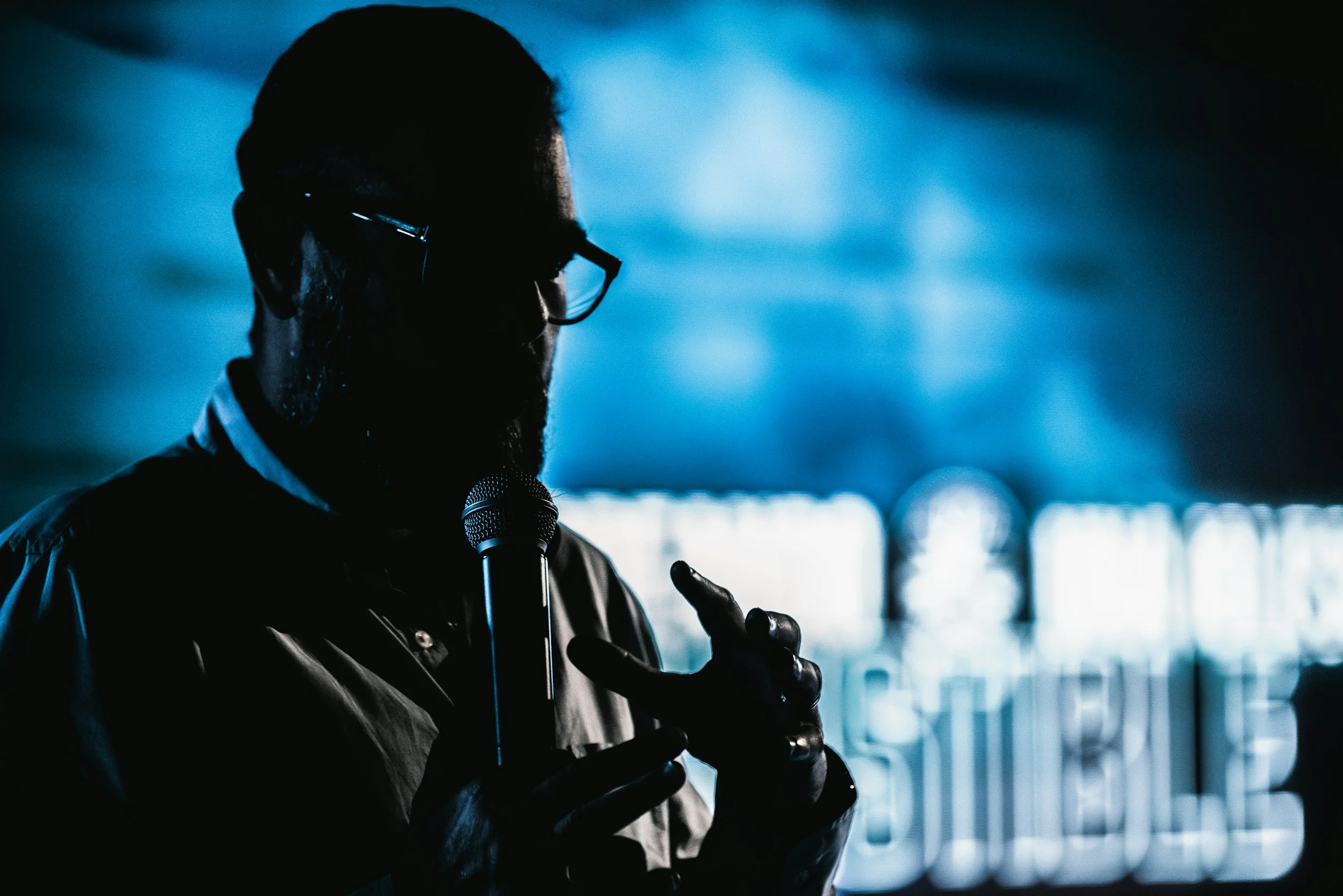 Photo of a man speaking. The man is in silhouette in front of a blue background with burred words on it.