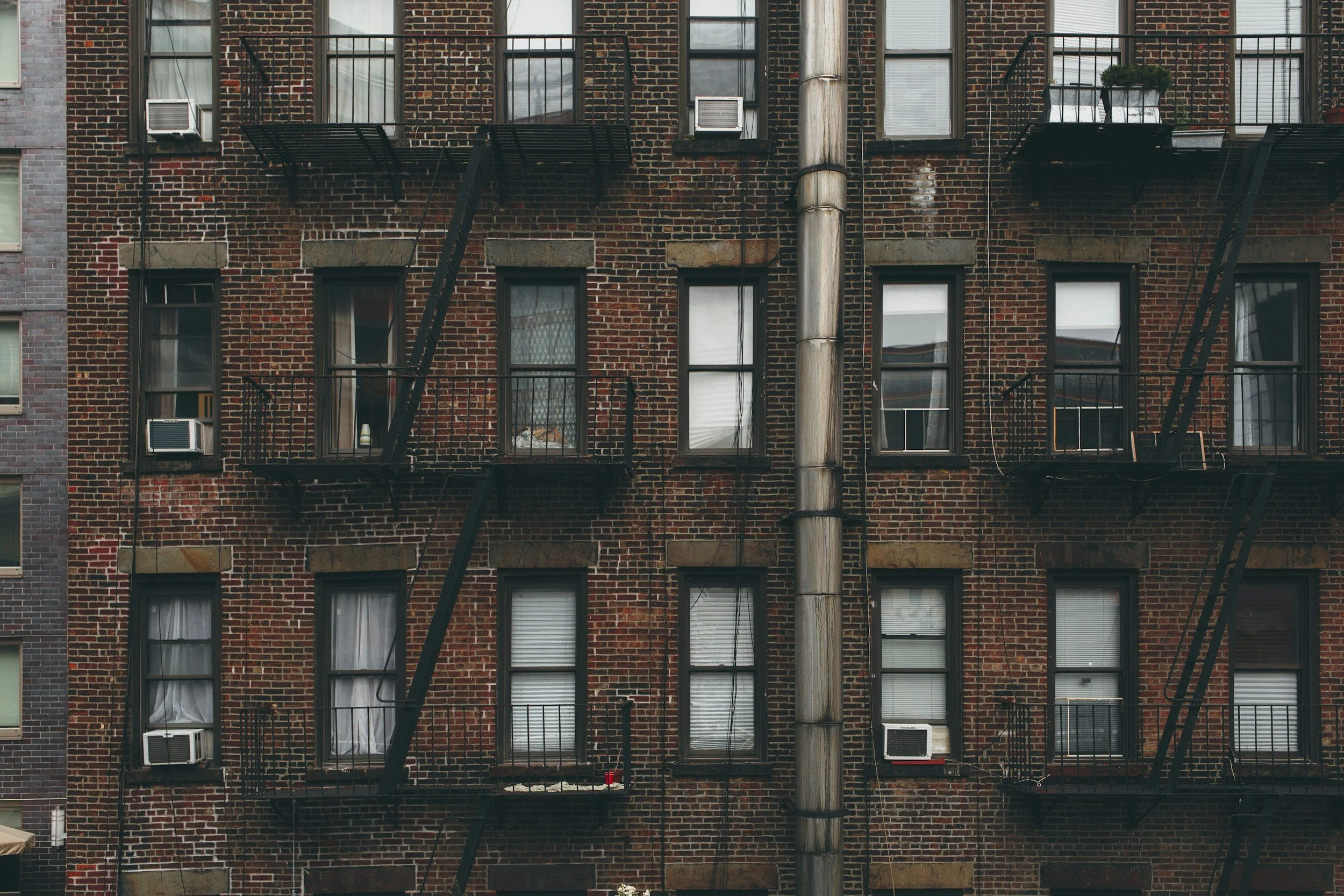 Image of a wall made of brick and windows with a fire escape and a pipe running up the side of a building