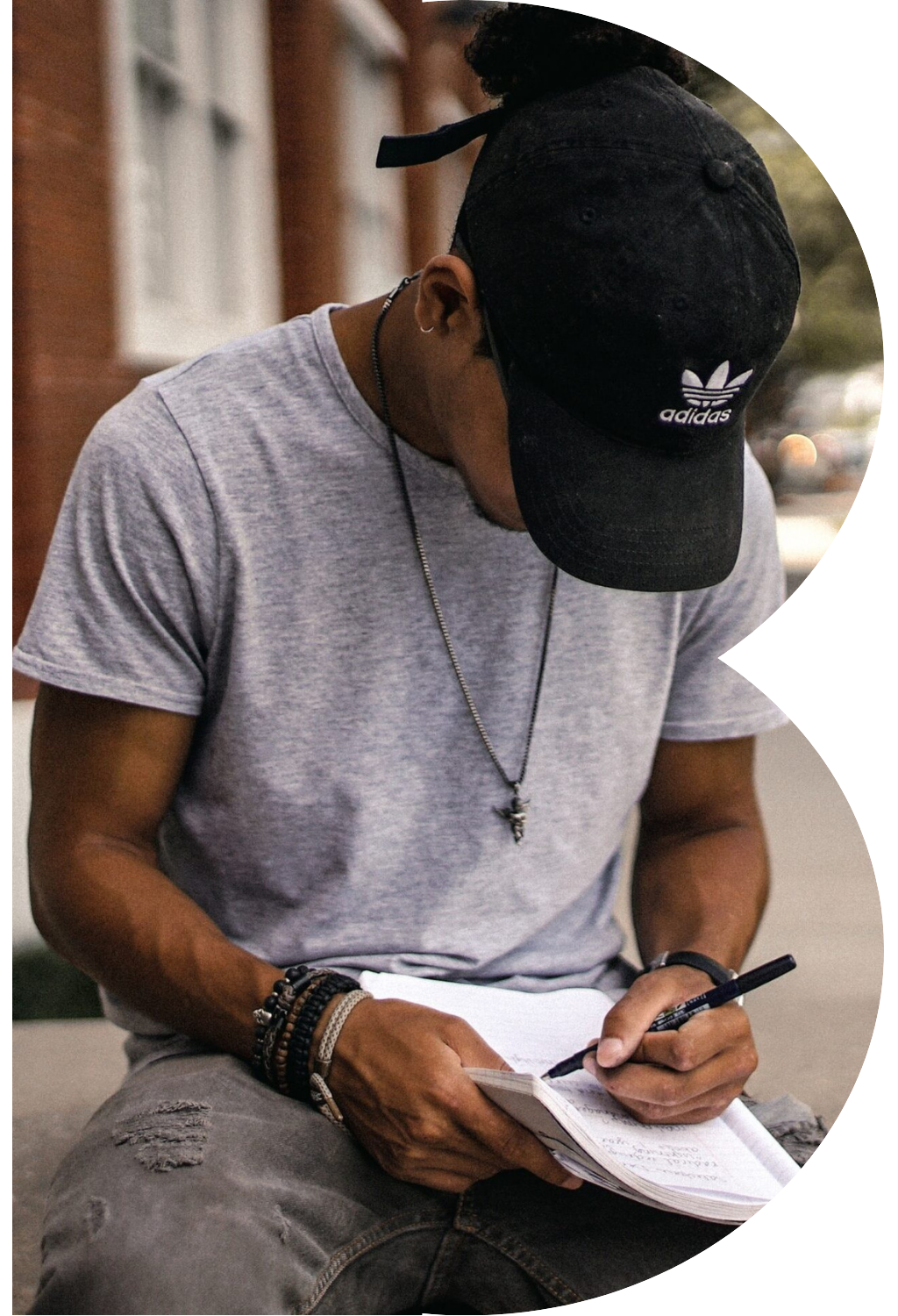 Photo of a young black man in a baseball hat sitting, looking down and writing in a notebook, in the shape of a capital B.