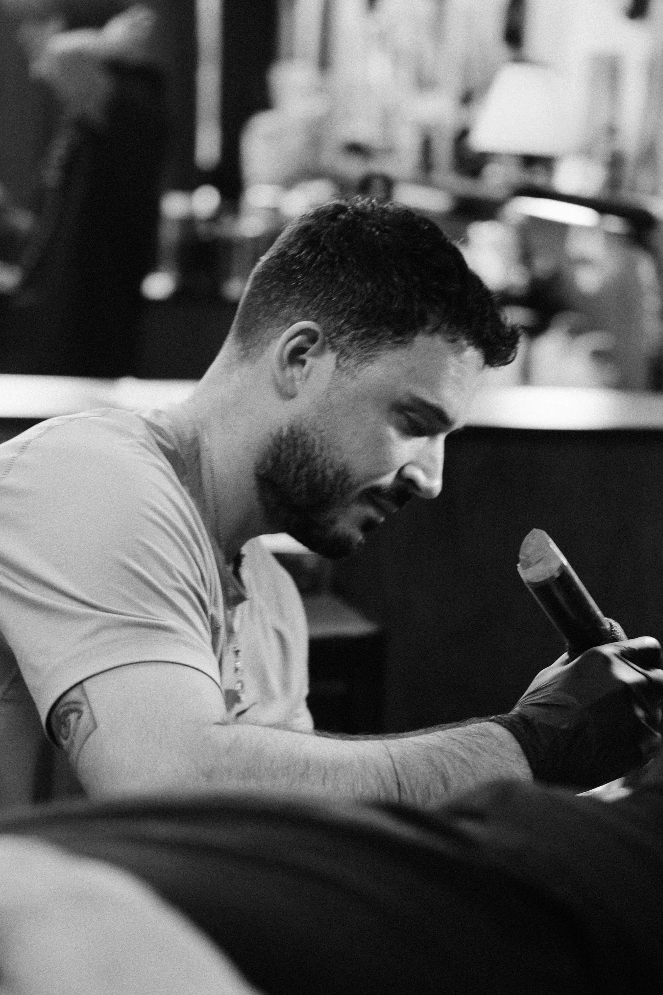 A young man with short dark hair and a beard, wearing a light-colored shirt and black gloves, is sitting and looking at his phone in a dimly lit indoor setting.