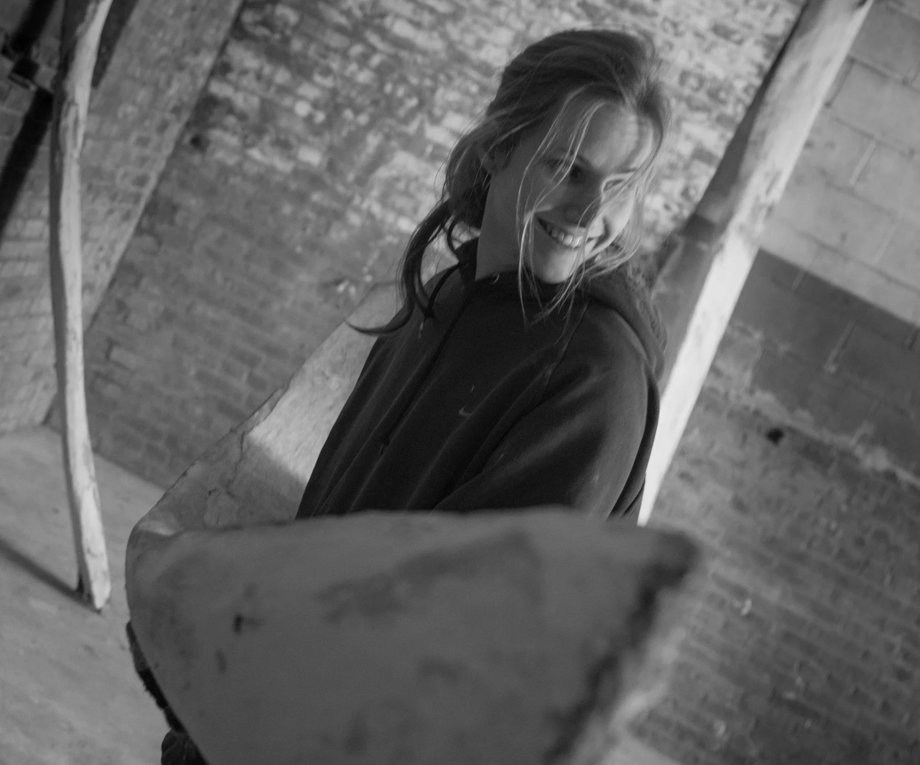 A young man with long hair smiling and reaching out towards the camera in an industrial-style brick and wood interior.