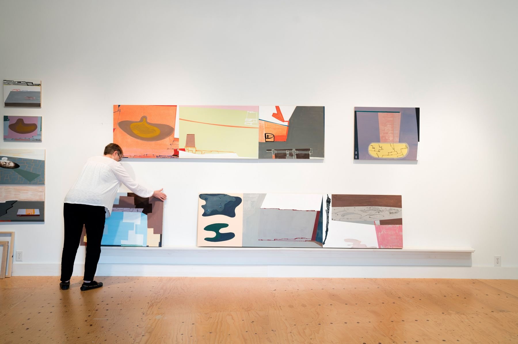 A person with glasses and wearing a white shirt and black pants is arranging or examining artwork in a gallery. The gallery wall displays multiple abstract paintings with varied colors and shapes, some mounted on the wall and others resting on a ledge below.