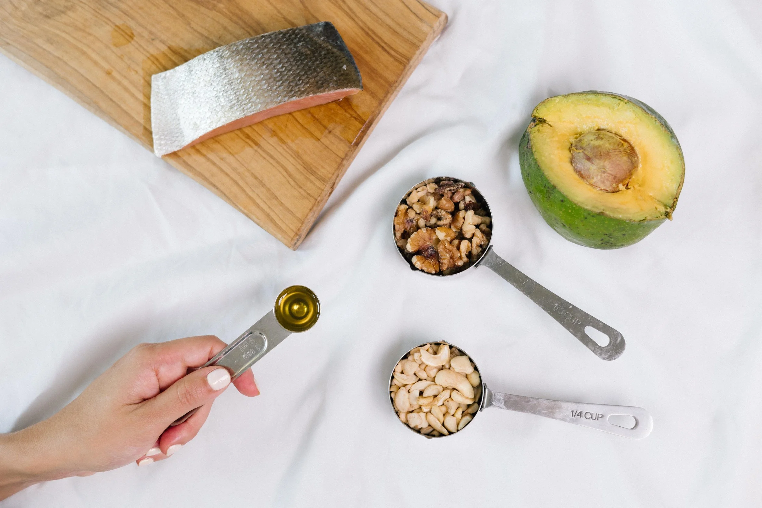 Top view of a white clothed table with scoops of nuts, an avocado, a wooden board with salmon on top, and a human hand with scoop of oil.