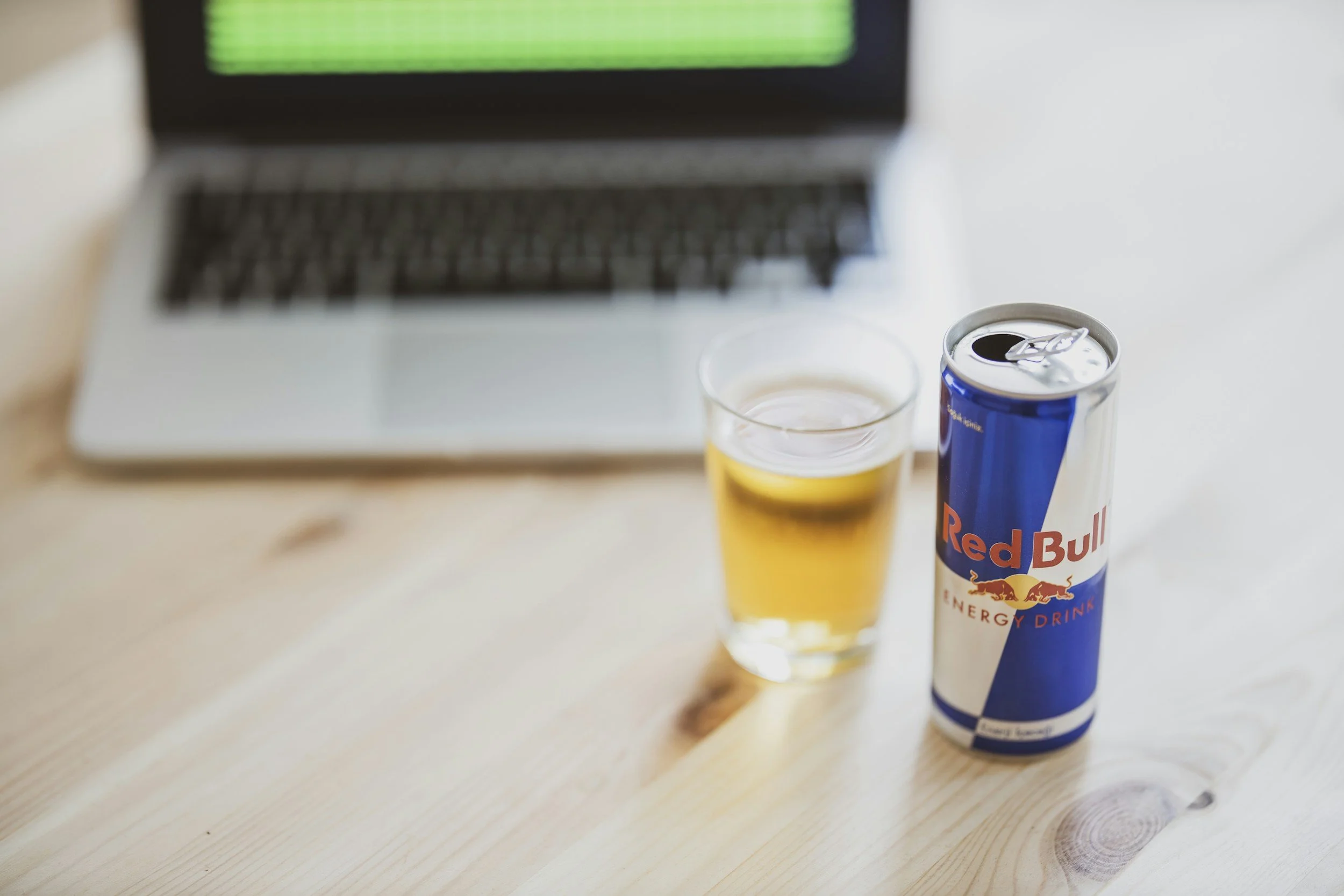 Can of Red Bull energy drink beside a glass filled with the beverage on a wooden table, with an open laptop blurred in the background.
