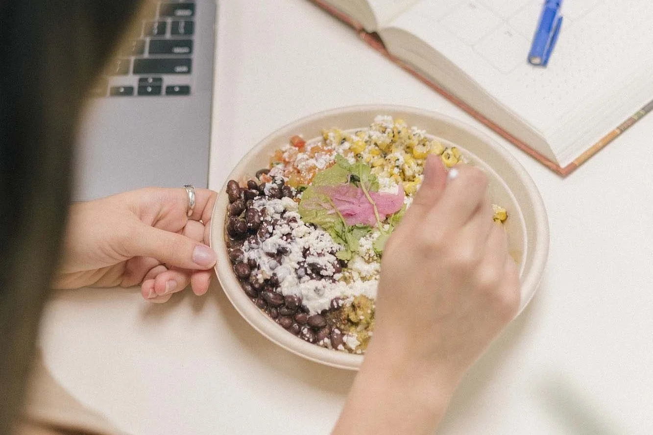Woman’s hands holding a carton bowl of nutritious food on an office desk, containing rice, beans, corn, tomatoes, and cheese.