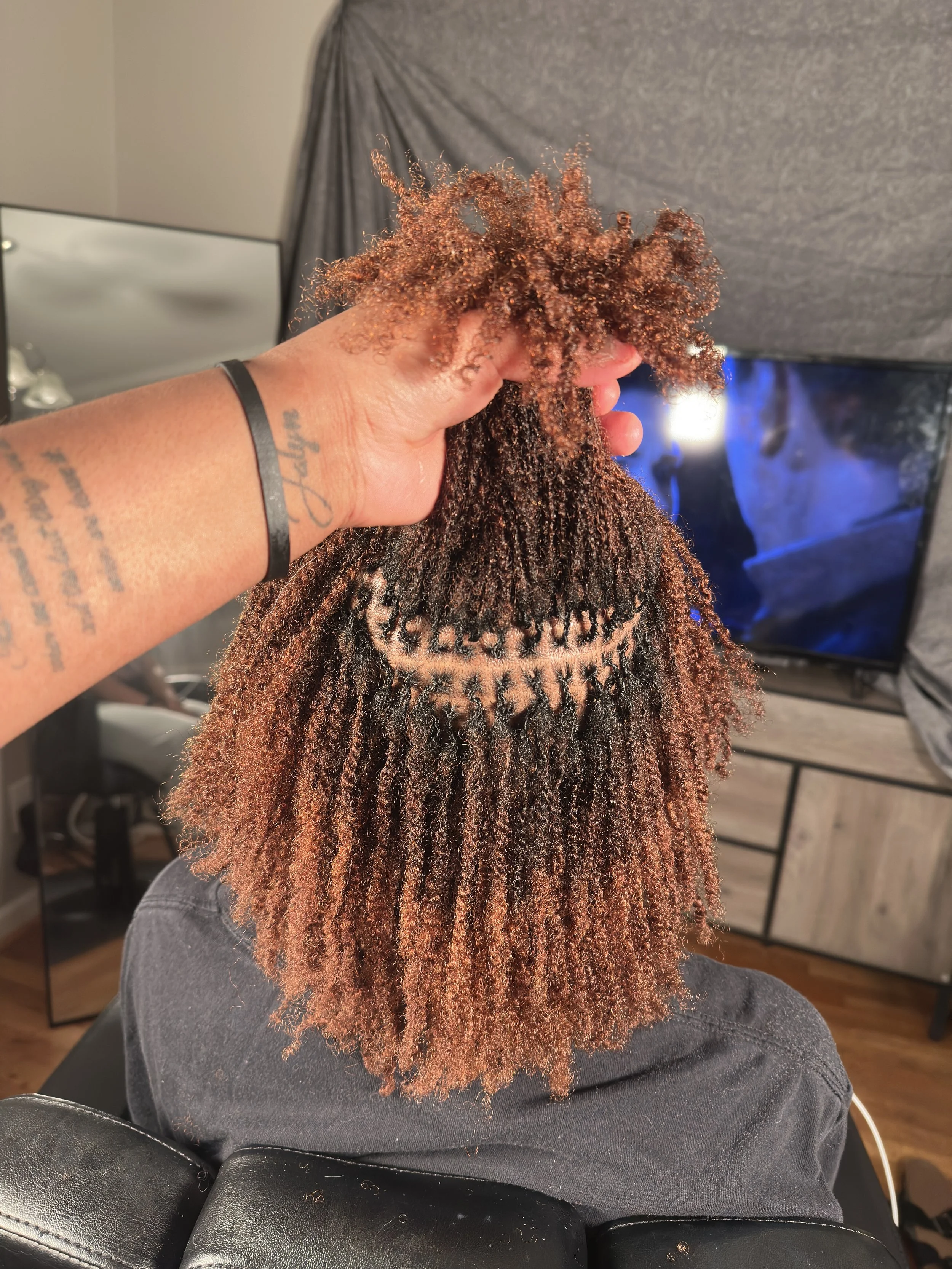 Close-up of a person's hand holding and examining the top of another person's head with textured, natural curly hair in a domestic kitchen setting.