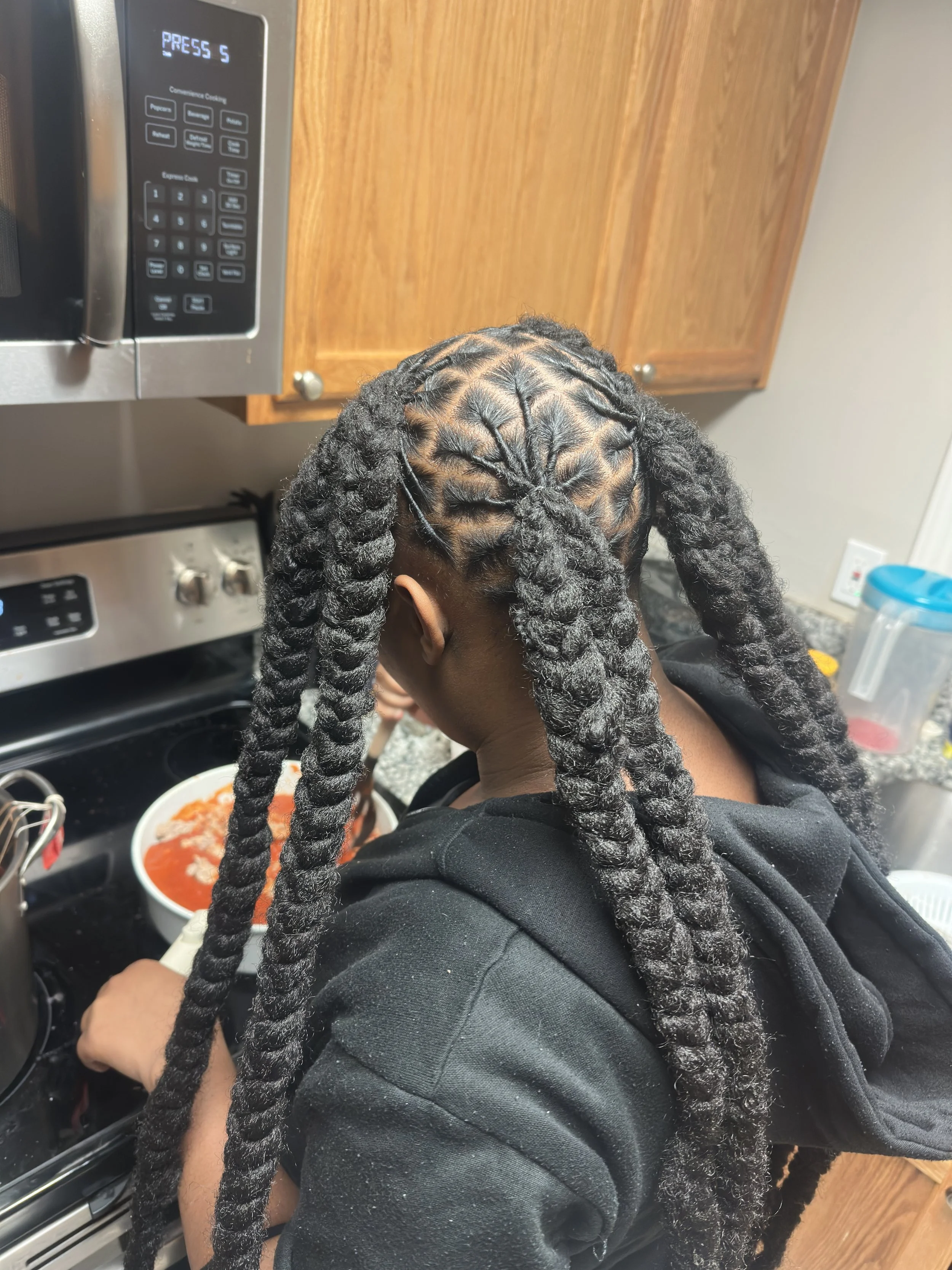 Person with styled, thick, twisted braids cooking in a kitchen with wooden cabinets and a microwave.