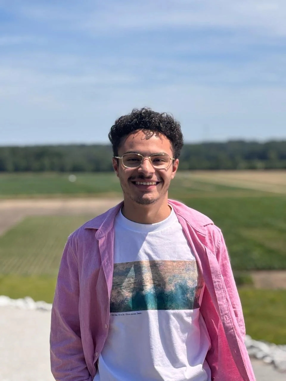 Young man with curly hair and glasses smiling, leaning against a tree outdoors during sunset.