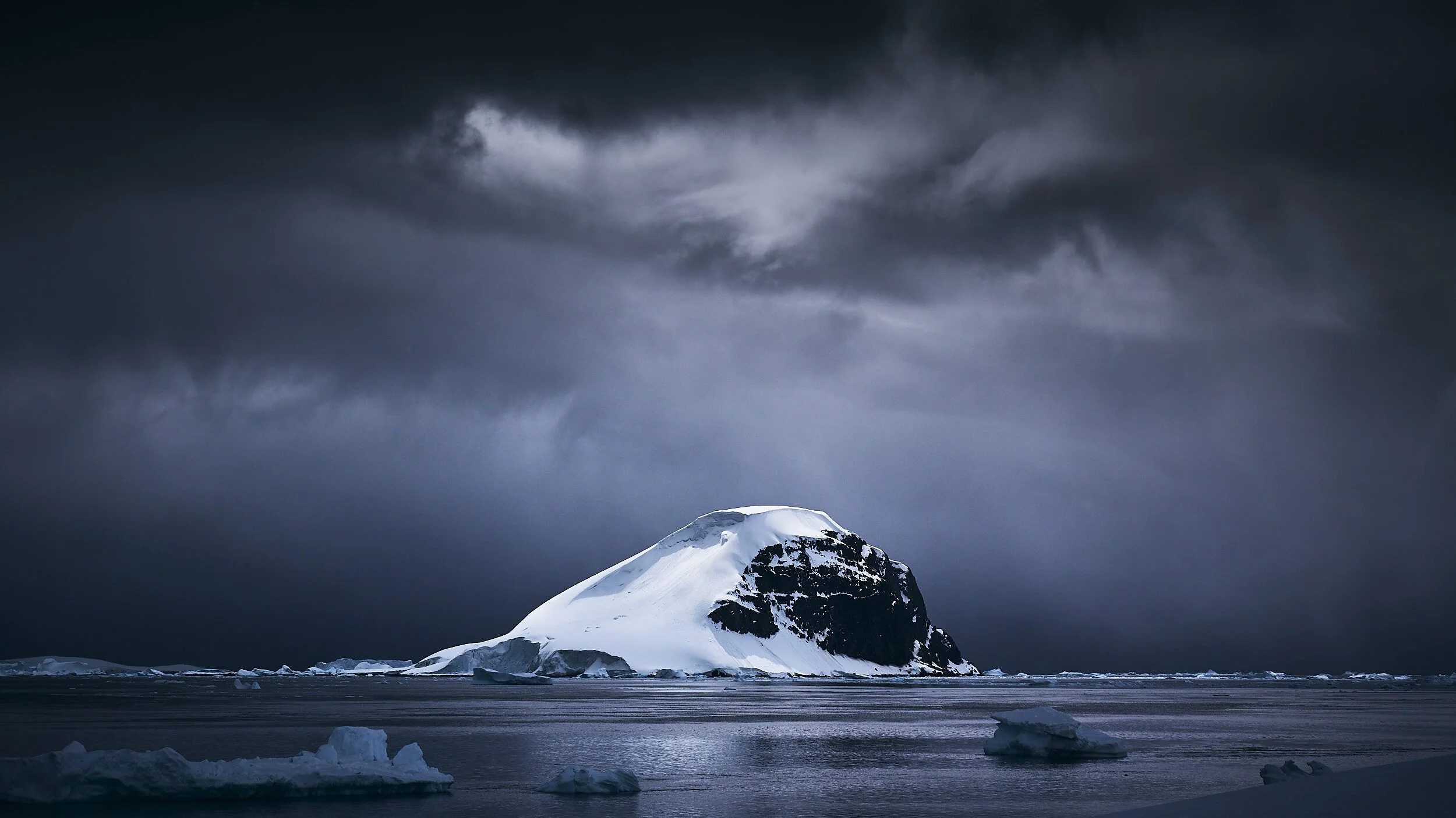 Snow-covered mountain peak under dark, clouded sky with floating ice chunks on the water's surface.