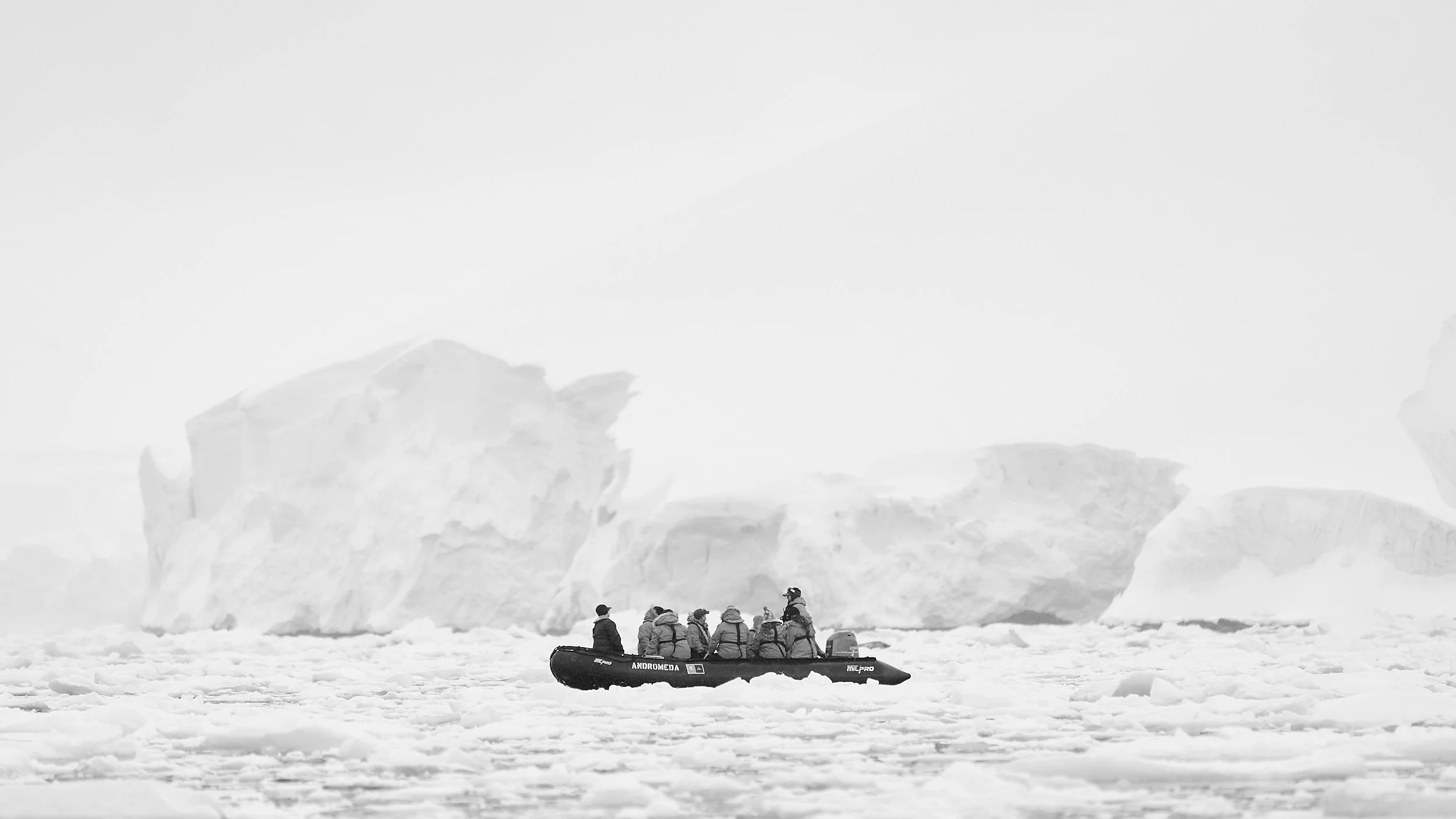 A group of people in winter clothing riding a small inflatable boat in icy waters, with large icebergs in the background.