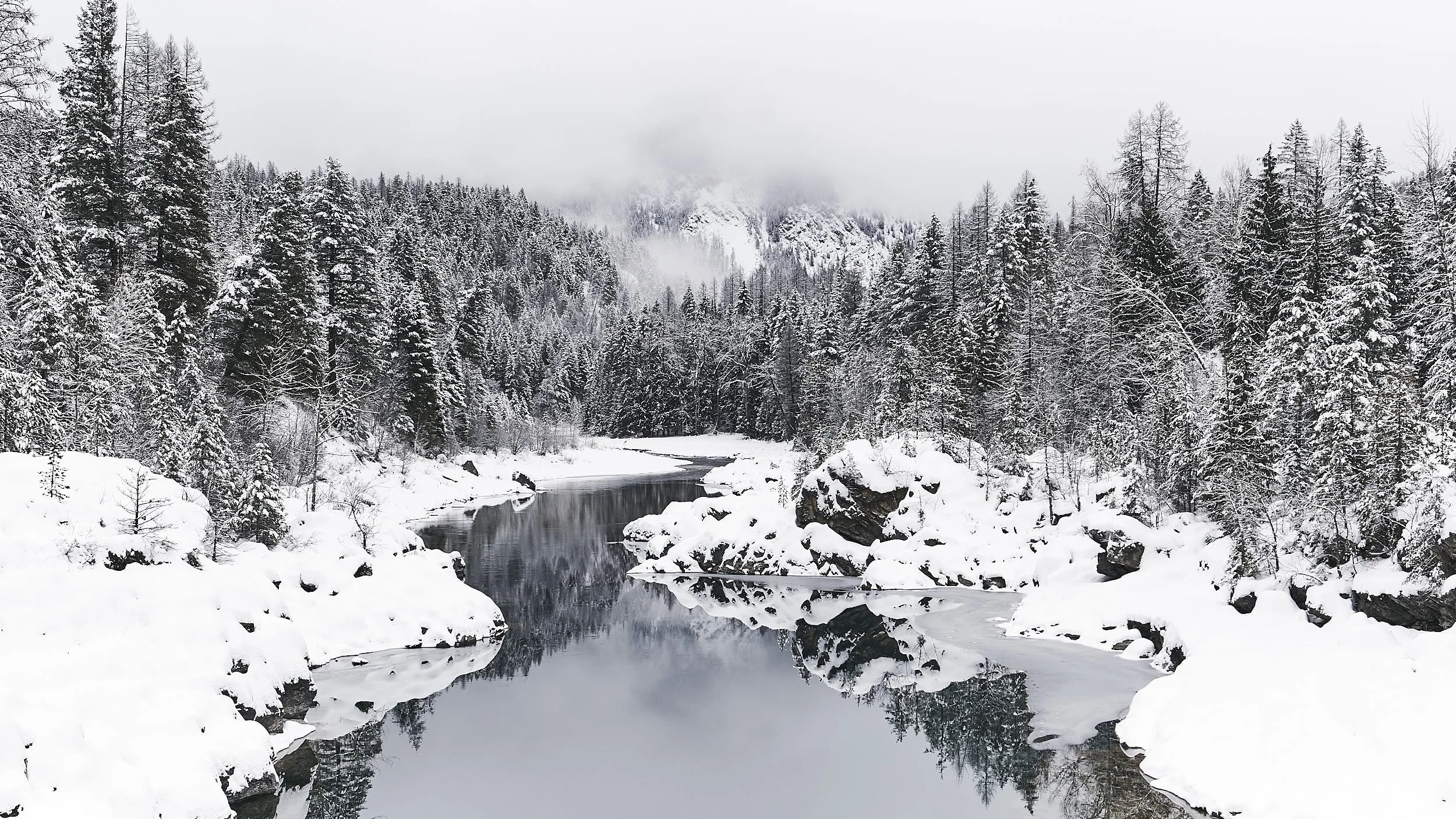 A snowy landscape featuring a river with partially frozen water, surrounded by snow-covered rocks and dense evergreen trees. Mountain peaks shrouded in clouds are visible in the background.