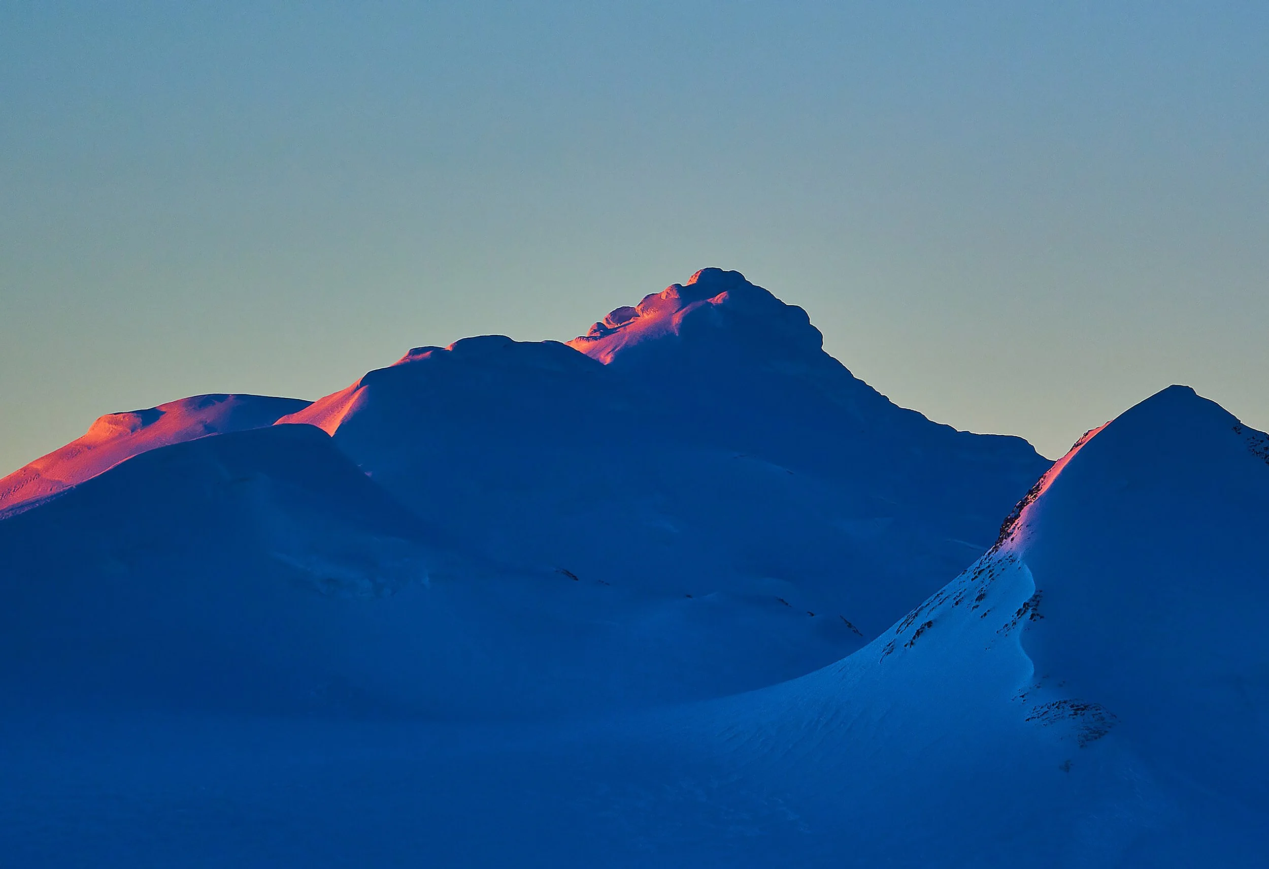 Snow-covered mountain peaks with a pinkish hue from the setting sun, against a clear blue sky.