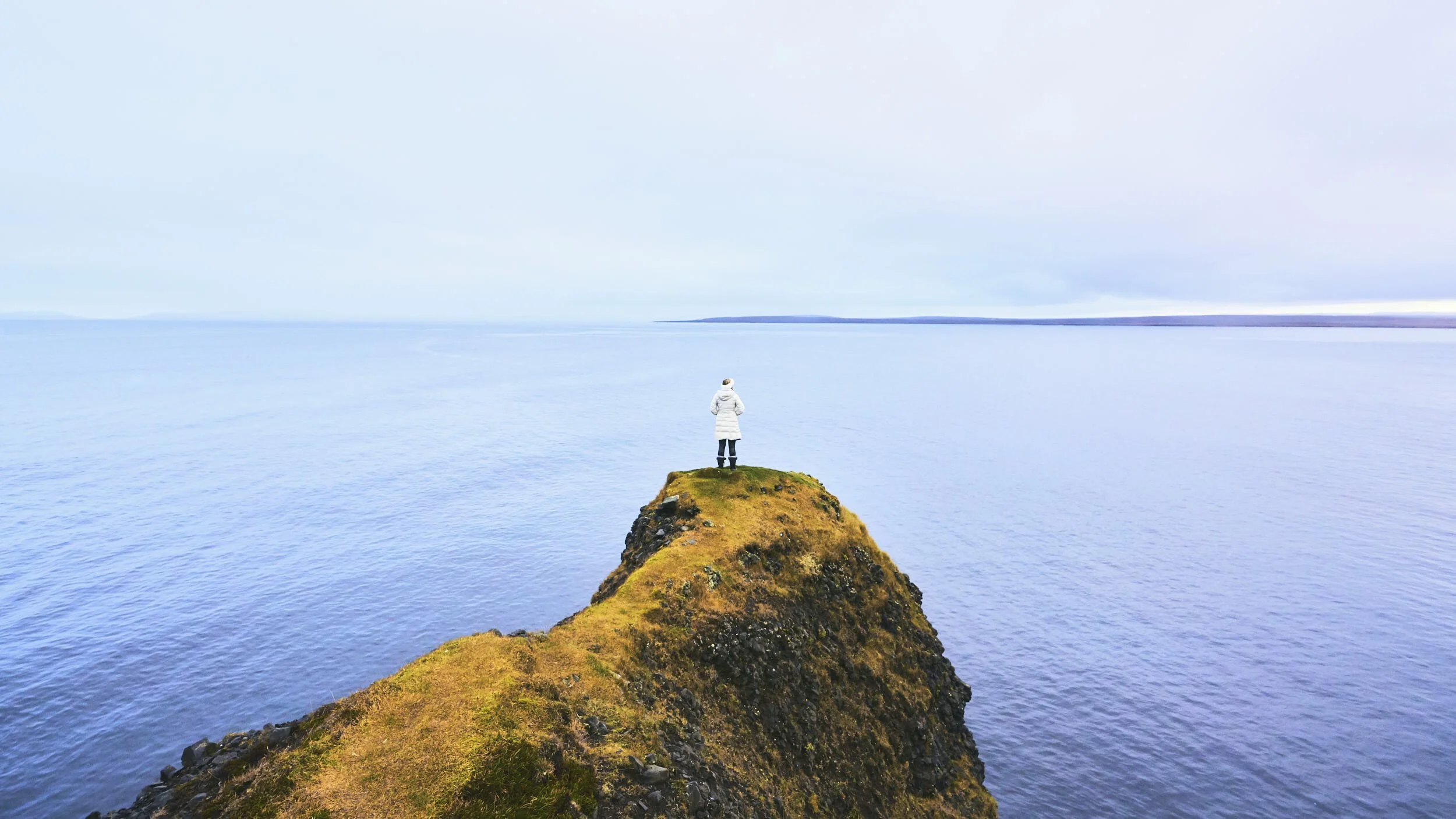 A person standing on a small grassy cliff overlooking a vast body of water with distant land on the horizon, under an overcast sky.
