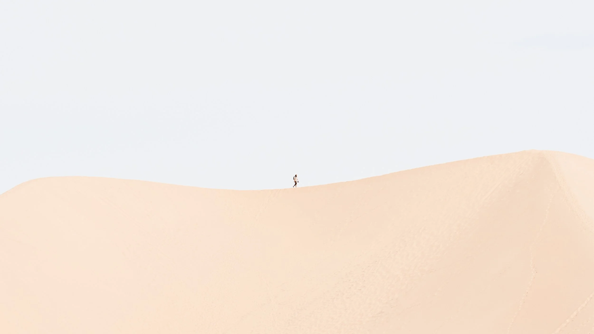 A person walking alone on a sand dune with a mostly clear sky in the background.